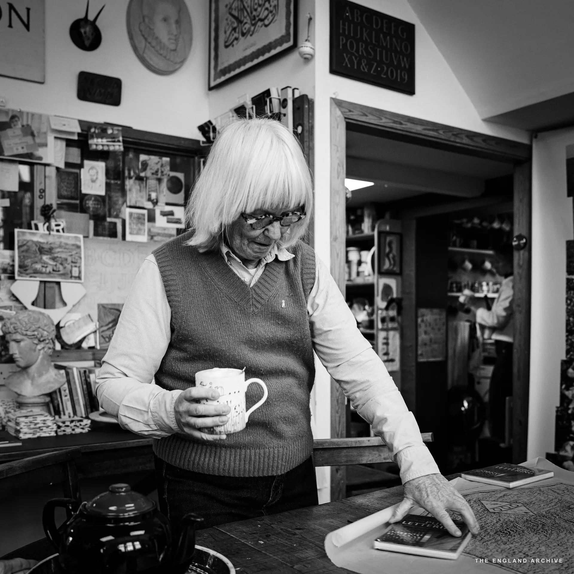 Lida Kindersley standing at the front-room counter in her sweater vest and white shirt, holding a floral mug, looking down at a book on the table. The workshop's pinboard wall covers the space behind her, the doorway through to the back workshop visible to the right.