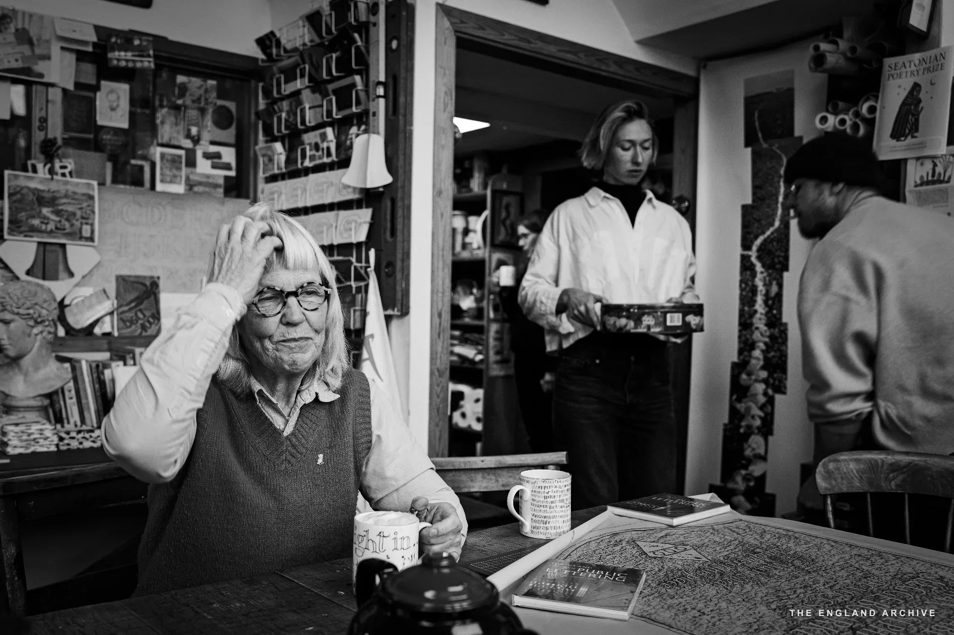 Lida Kindersley seated in the foreground, hand to her head, eyes turned away in a pause; behind her in the doorway Roxanne Kindersley stands in a white shirt holding a small object, and a man in a cream jumper turns to one side - three generations of the workshop in the same frame.