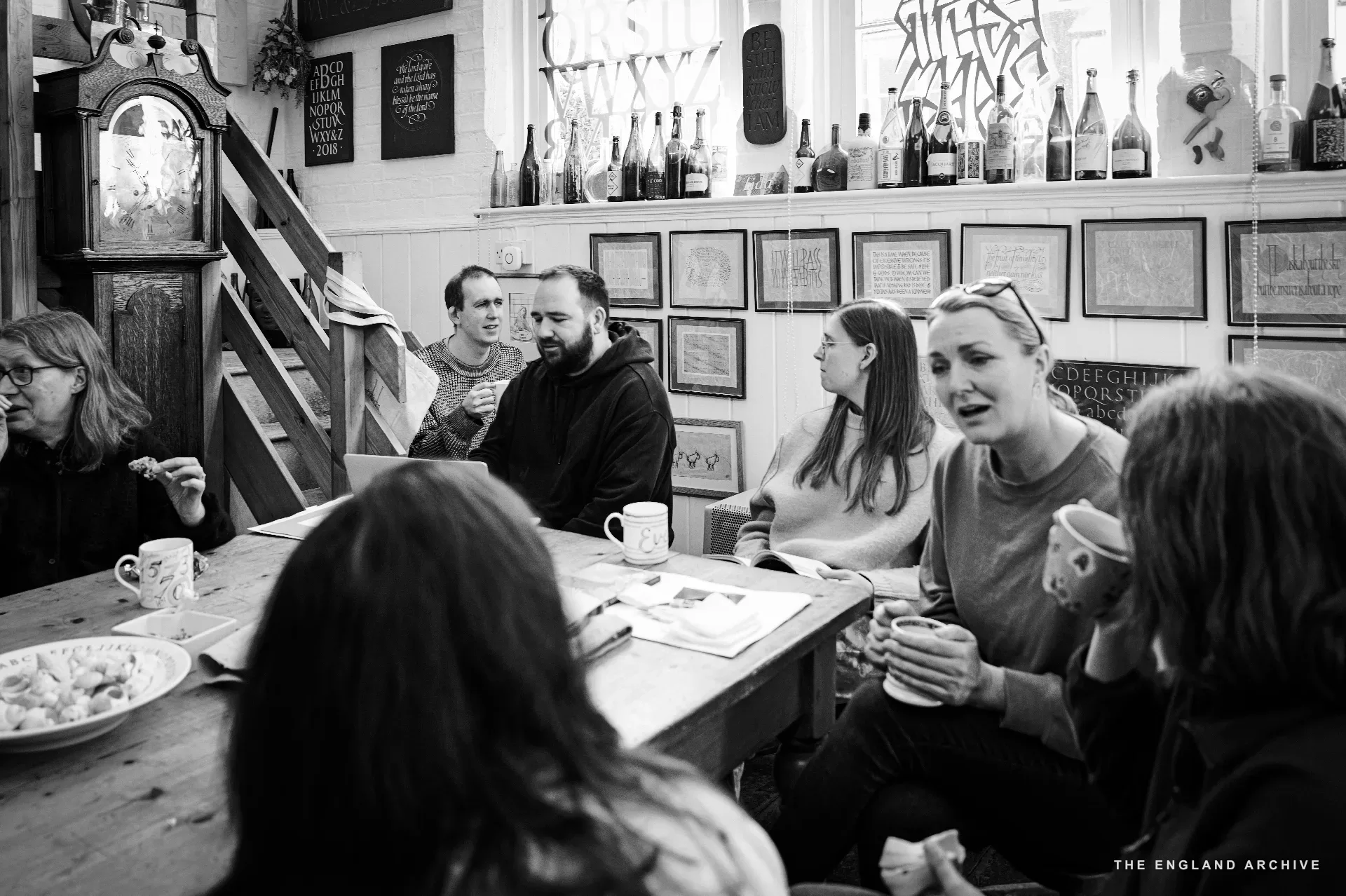 A wide view of the front-room table mid-tea: six or seven team members and visitors gathered around it in conversation, the longcase clock and painted carved sign visible on the wall, the high shelf above lined with bottles.