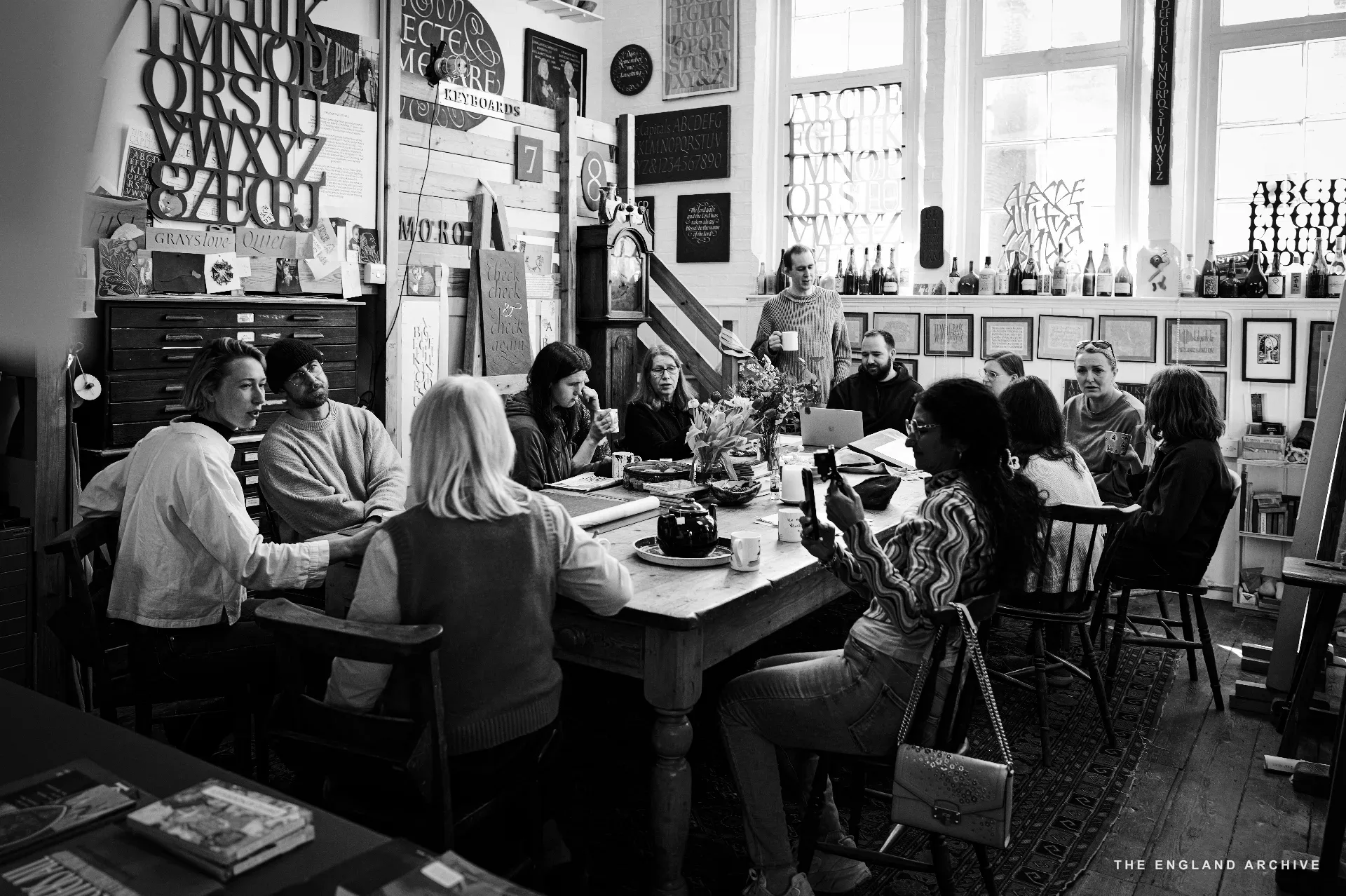 A wide environmental view of the front room: the big table at centre with team members seated around it, Lida's position to the left, the printer's tray cabinet visible behind, the painted sign and longcase clock to the back, framed letter samples covering the wall on the right.