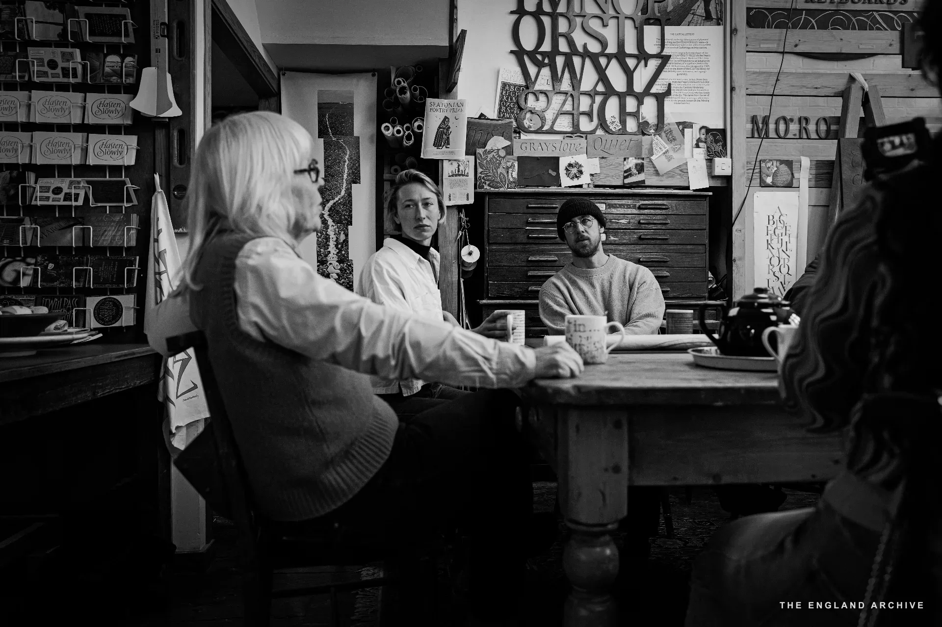 Three figures around the front-room table: Lida Kindersley to the left in her vest and pale shirt, a young woman in the centre, a man in a beanie on the right holding a mug. Behind them the workshop's painted carved 'MORO' sign and the printer's tray cabinet.