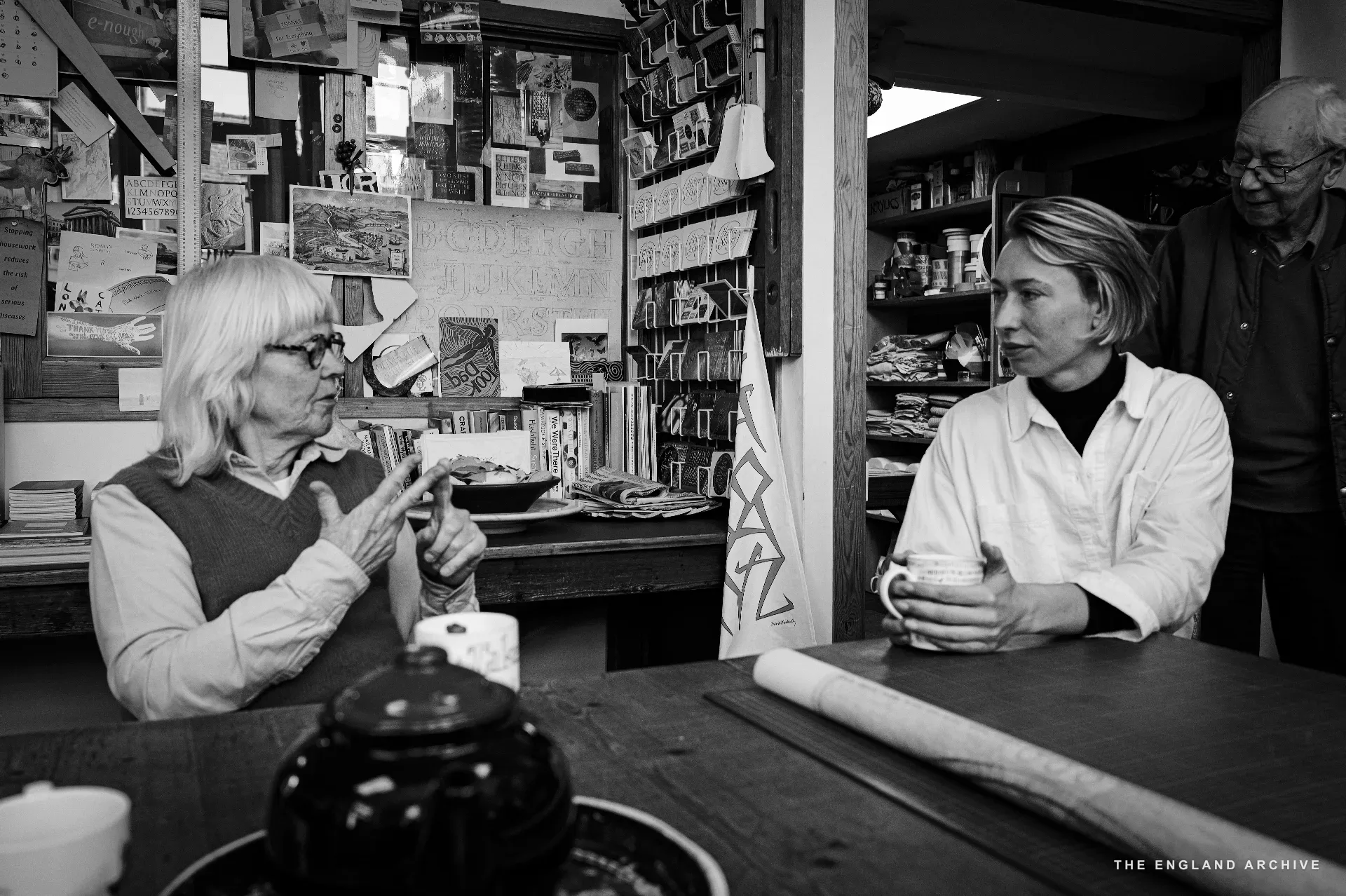 Lida Kindersley to the left in profile, talking, hands raised in mid-explanation; Roxanne Kindersley to the right in a white shirt, listening, holding a mug. A figure in the back of the workshop is just visible. The table between them holds a teapot and a long roll of paper.