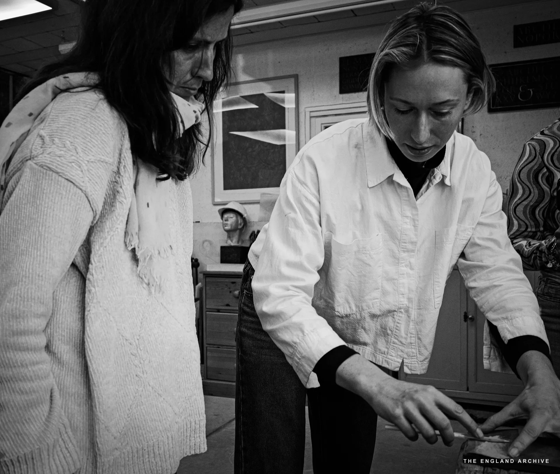 Roxanne Kindersley to the right in a white shirt, leaning over a bench and showing a slate panel; the new apprentice - the young woman who had walked in off the street that morning - watches from the left in a knitted cream cardigan, her dark hair falling forward. A framed image hangs on the wall behind.