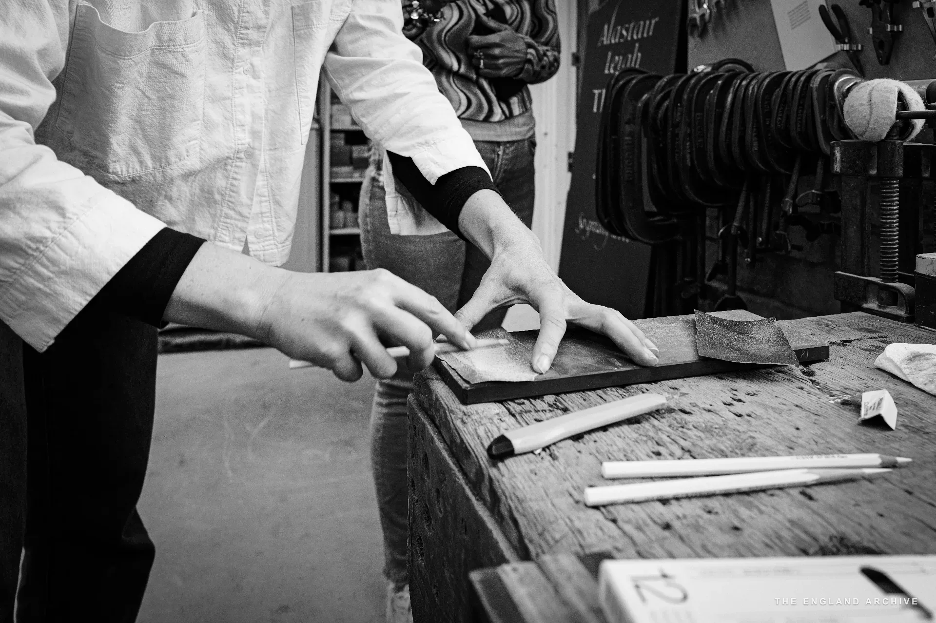 Close on Roxanne Kindersley's hands at the bench, fingertips resting on a small slate panel showing the workshop's setting-out marks; a sharpened pencil and another pencil rest on the wood beside it. The workshop's tool clamps are racked along the bench edge to the right.