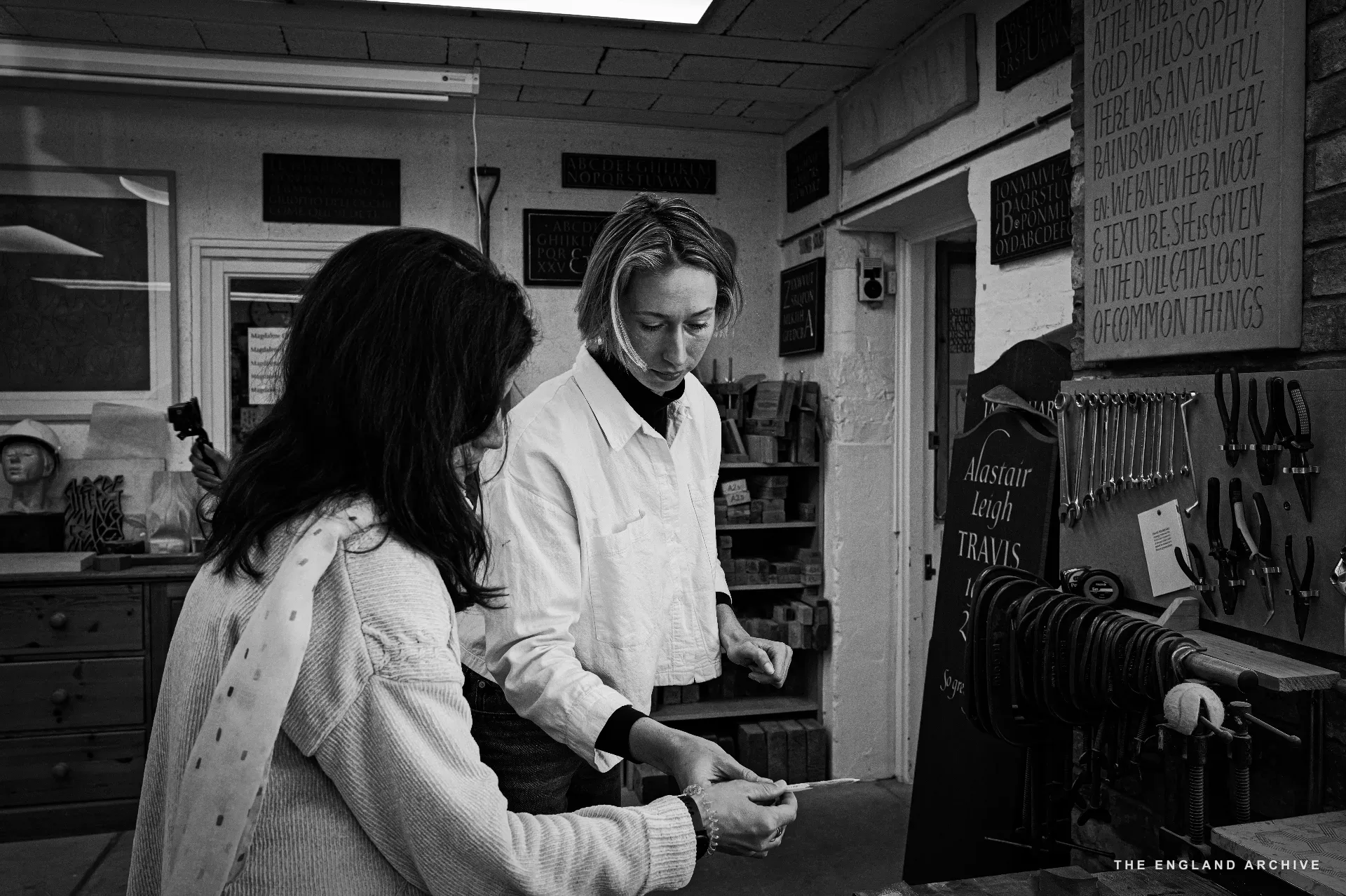A workshop scene: Roxanne Kindersley standing at a bench in a white shirt, examining a small object held in her hands; a woman in a cream cardigan to the left watching closely. The workshop's tool wall fills the right side - chisels, mallets, and a TRAVIS sign.