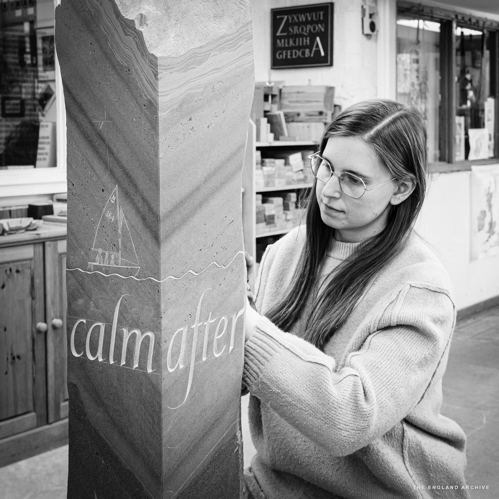 Emily looking sideways at the tall slate pillar she is carving, her hands resting on it. The visible side of the stone is cut with a sailing boat and the words 'calm after' in flowing italic capitals. Behind her, the workshop's alphabet panel and the bookshop visible through the window.