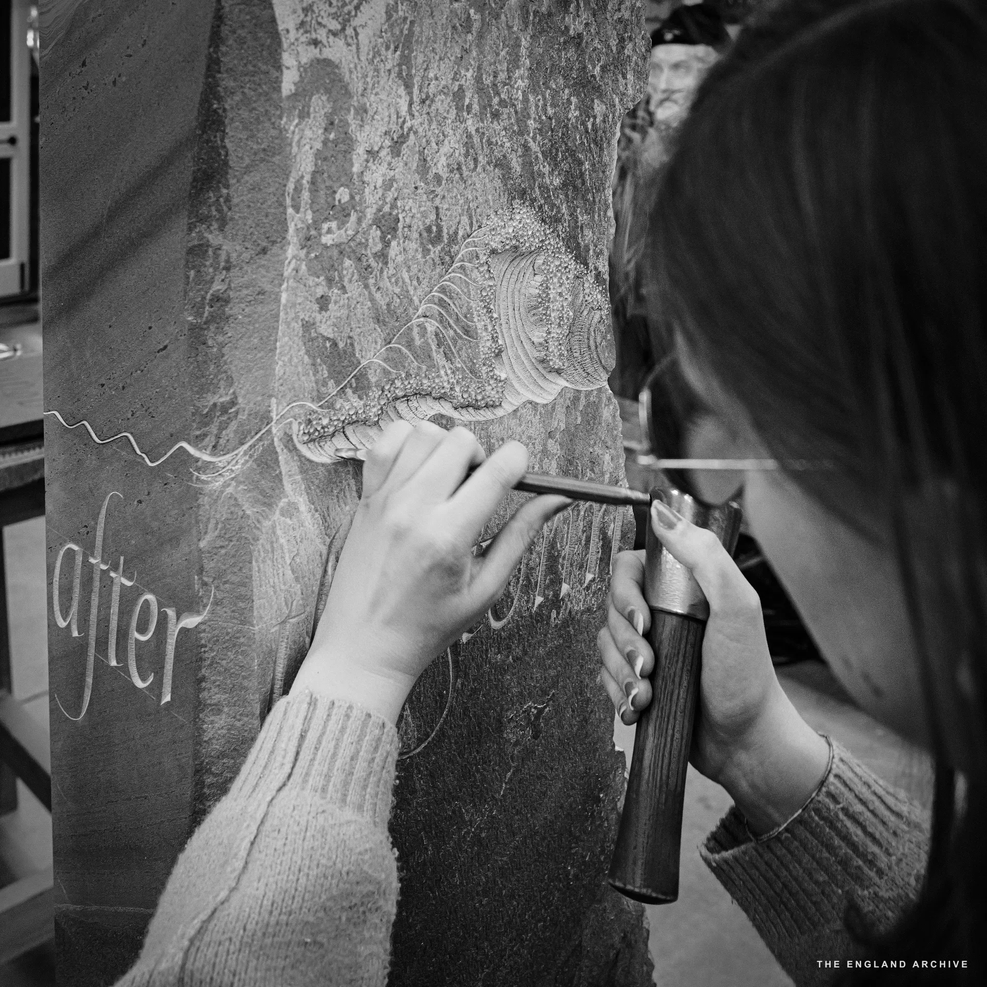 Close on Emily's hands at work on the pillar: her left hand steadying the chisel, right hand holding the dummy, carving the wave detail above the words 'after'. Her dark hair falls forward across her face as she leans into the stone.