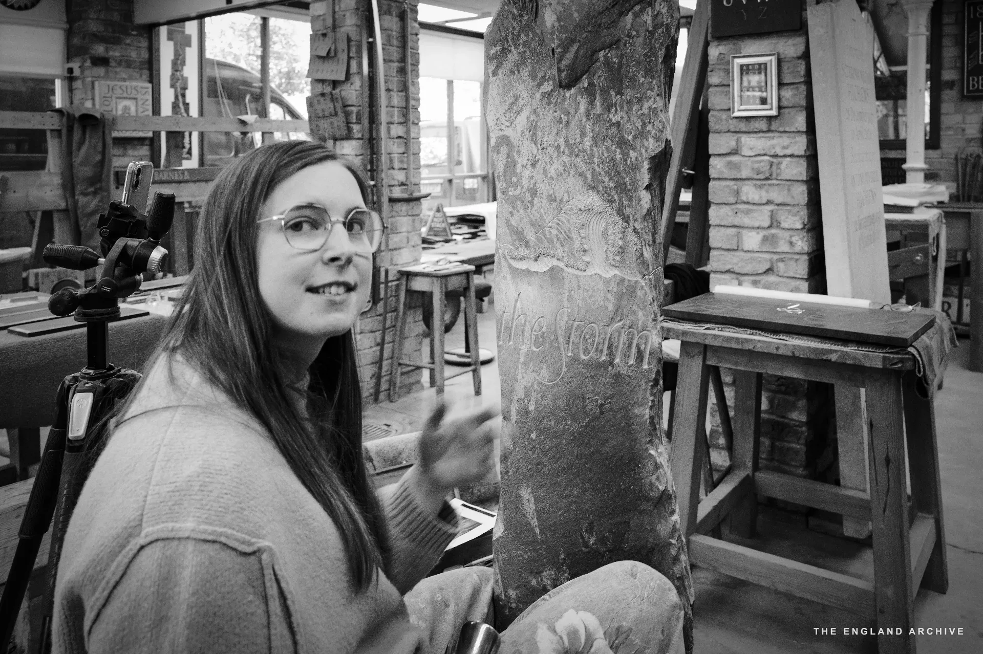 Emily with her chisel held loosely, looking back over her shoulder at the camera with a faint smile, beside the tall slate pillar she is carving. The back workshop and a tripod-mounted camera visible behind her.
