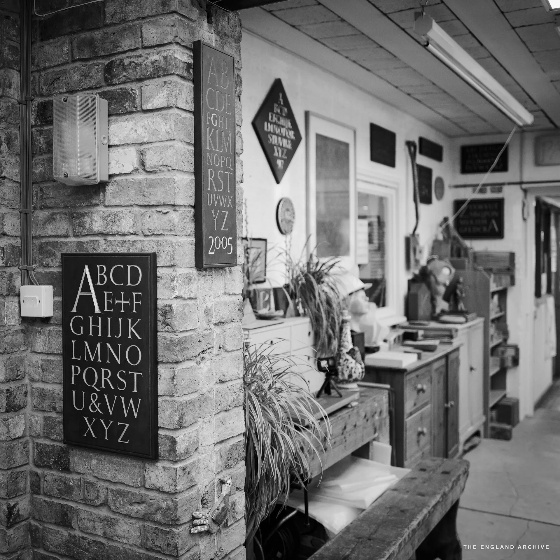 The corridor running between the front room and the main workshop, exposed brick column on the left mounted with a hand-cut alphabet panel and a slate inscription dated 2005, the back workshop visible through an opening, a desk with a figure working at it.