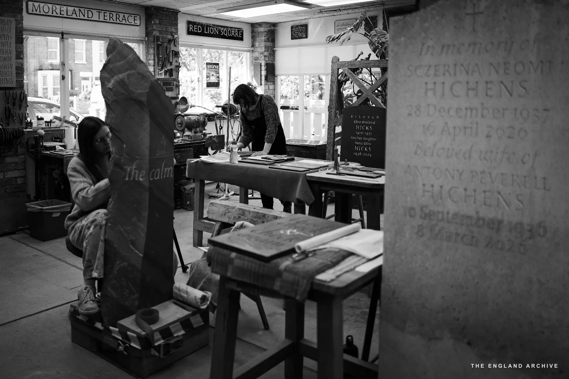 A wide view through the workshop: Emily at her tall pillar in profile to the left, the back room's benches and figures working visible through the doorway, a slate tomb panel inscribed for 'Sczerina Neomi Hichens' propped to the right of frame.