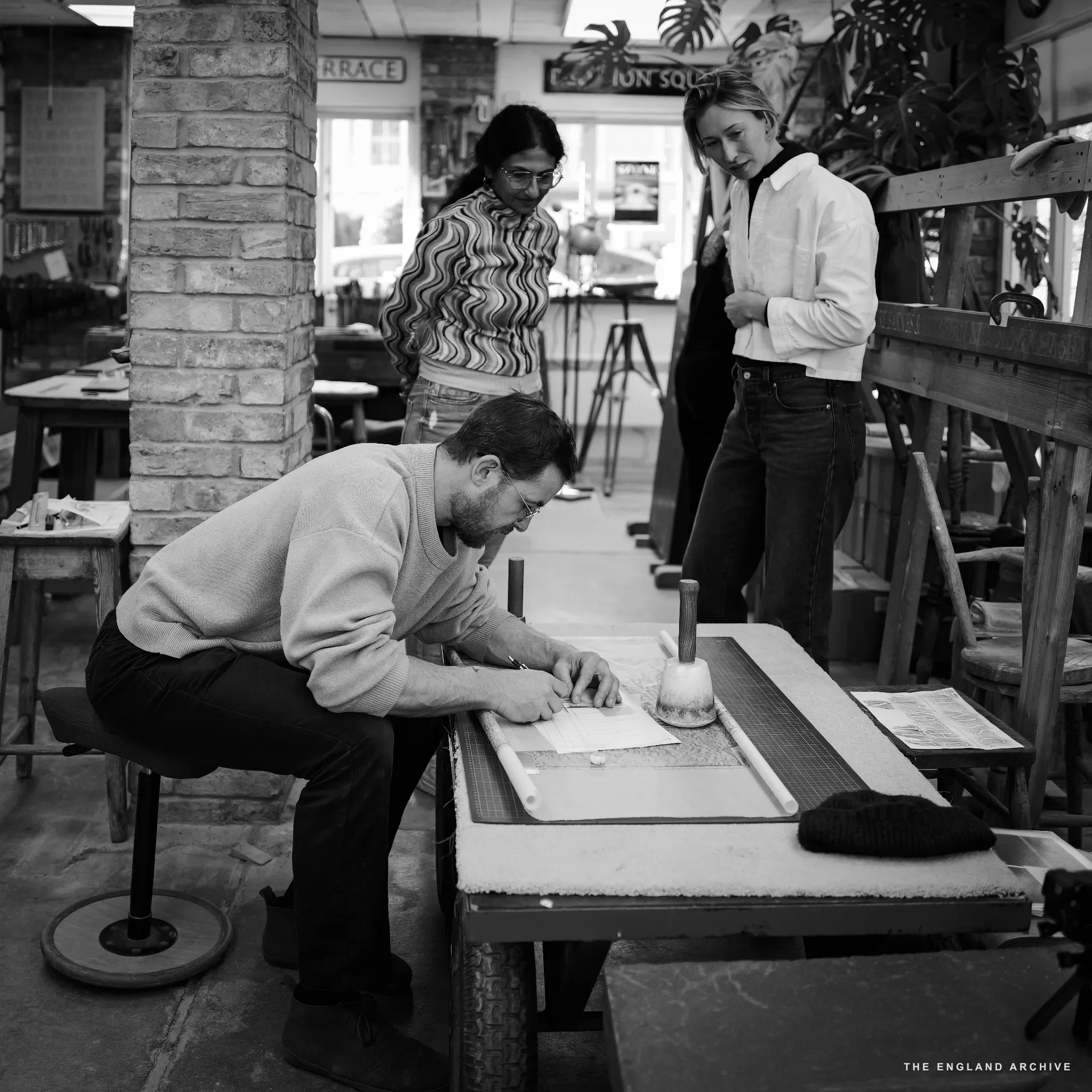 A bearded man in a pale jumper and glasses sitting low at the bench, marking out a stone with a pencil; behind him two figures stand watching - a woman in a striped top and Roxanne Kindersley in a white shirt - with the workshop's plants and brick column behind.