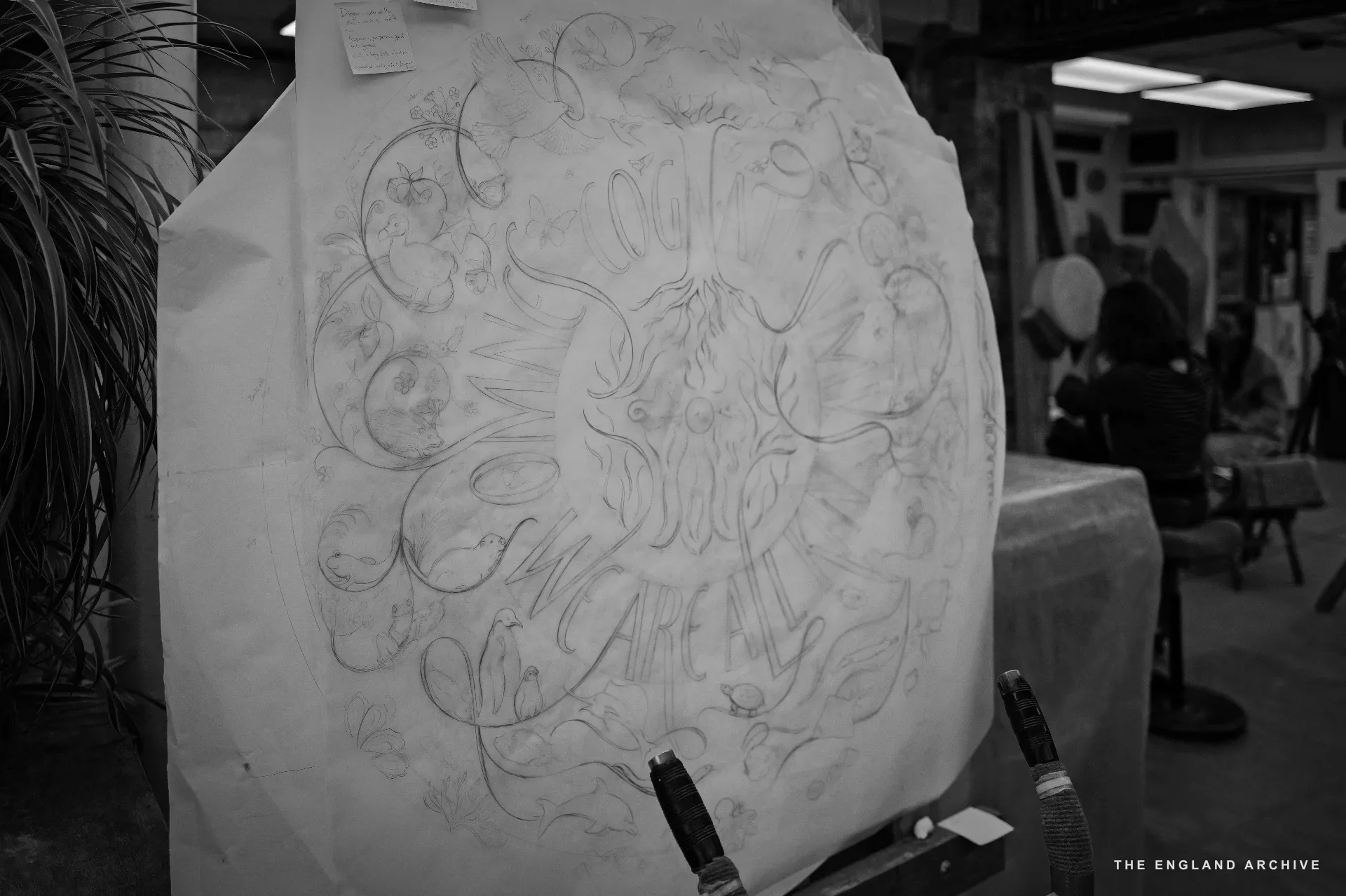 A pencil setting-out drawing on white paper, mounted to a vertical easel: an intricate circular design of swooping vines, leaves, and the lettering 'CALM CALM AFTER' woven through the curves - the working sketch for the pillar Emily is carving.