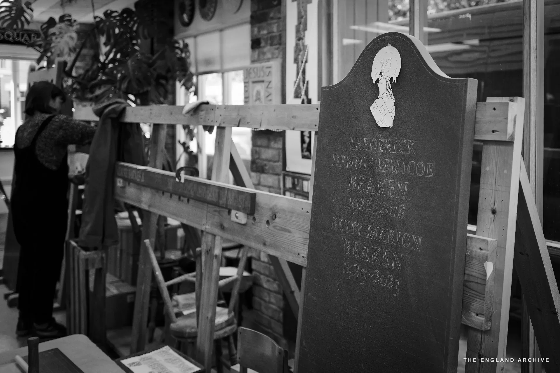 A finished slate memorial in the workshop, foreground right: dark stone with the inscription 'FREDERICK DENNIS JELLICOE BEAKEN 1936-2018 / BETTY MARION BEAKEN 1939-2023'. In the soft-focus background a workshop member moves between benches.