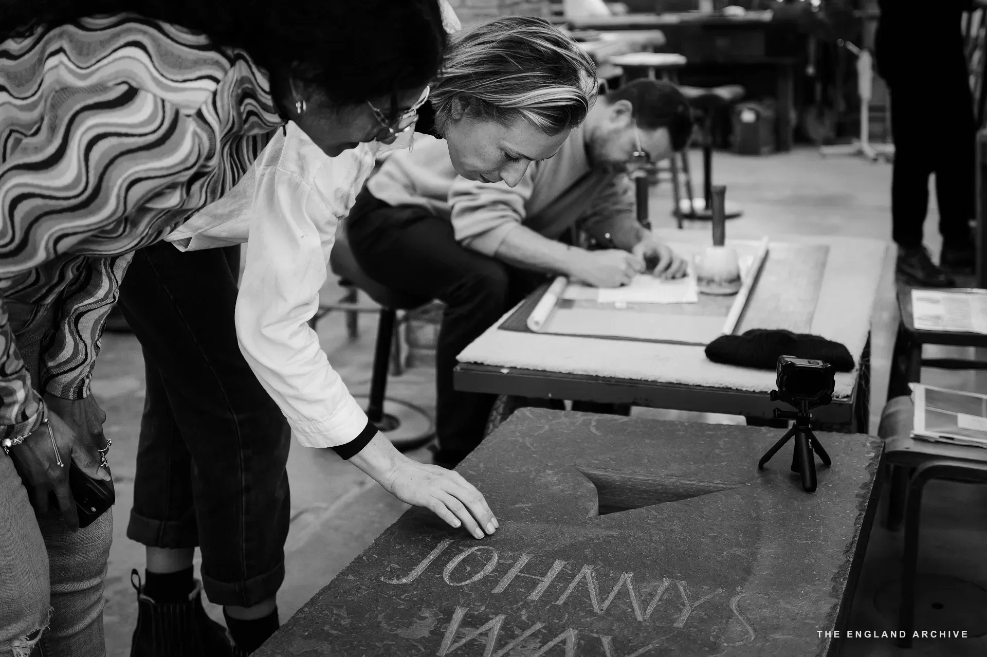 A scene at the bench: a woman in a wave-patterned top to the left examining a slate panel cut with the words 'JOHNNY'S WAY', Roxanne Kindersley centre-right leaning in to look at it, another figure beyond her bent to the work. The workshop's tool rack visible to the right.