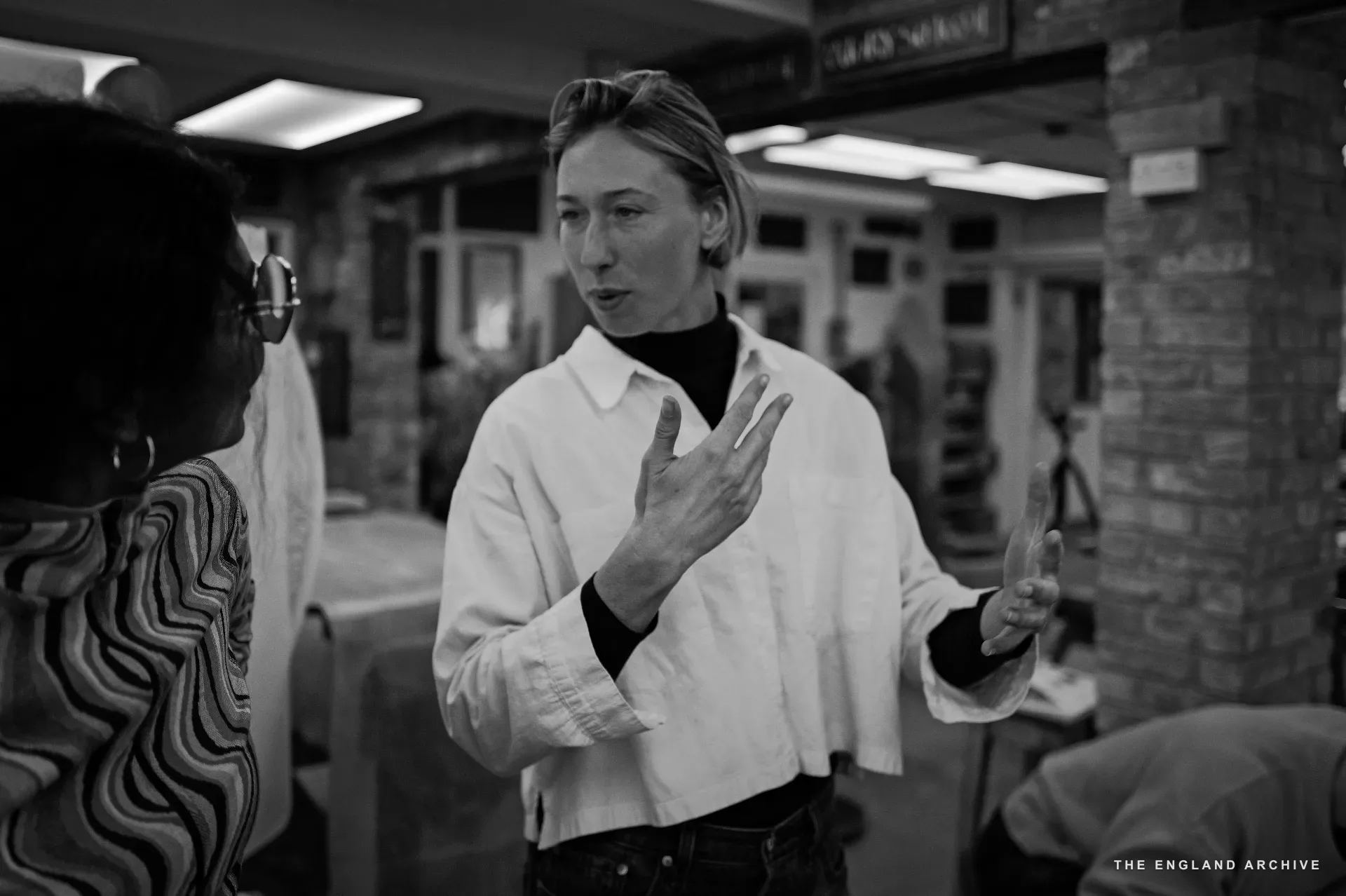 Roxanne Kindersley in profile in the Cardozo Kindersley Workshop, hands raised mid-explanation, in a white shirt over a black turtleneck.