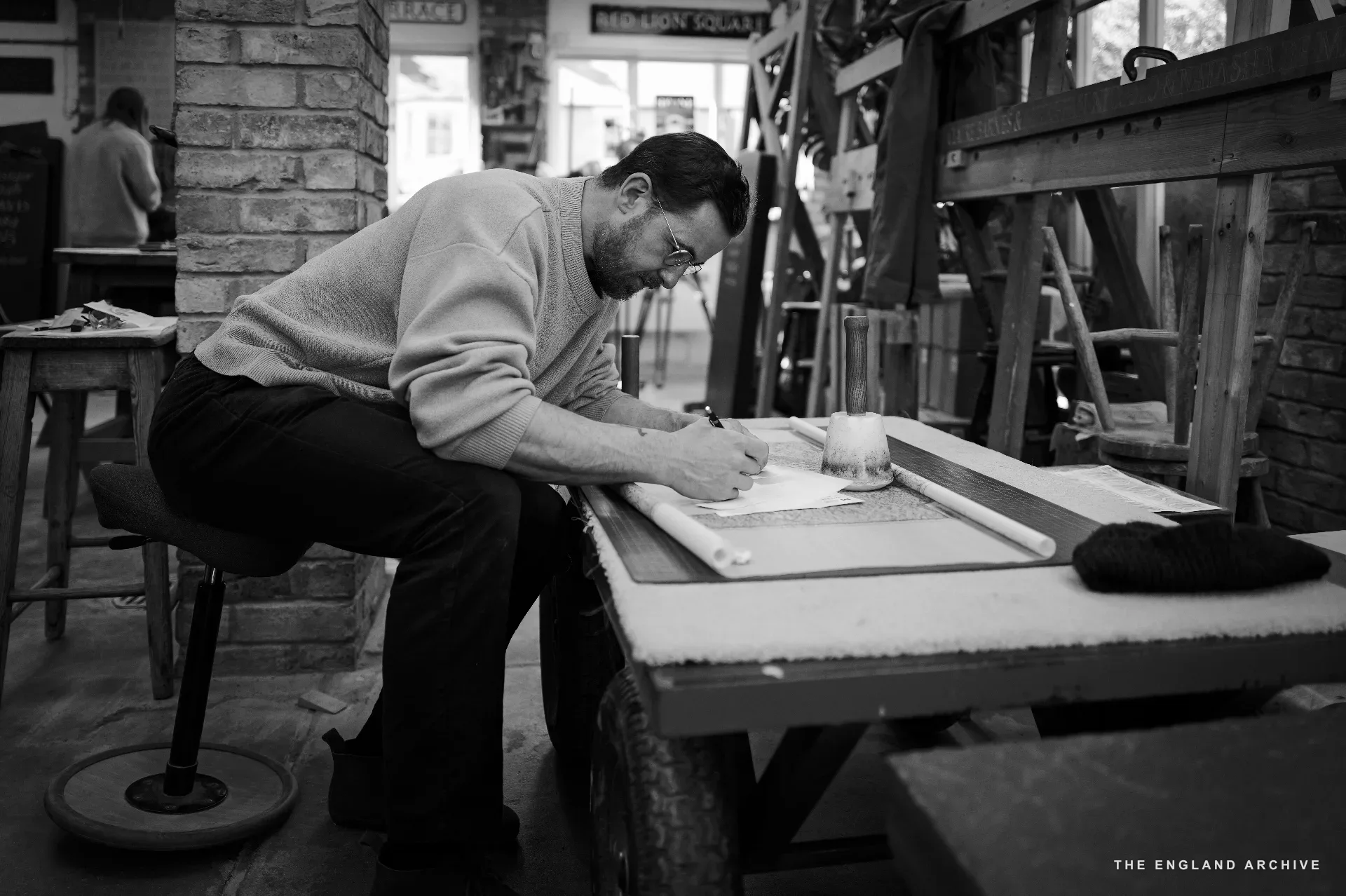 A bearded man in a pale jumper and glasses leaning over a stone laid flat on a wooden bench, working alone at the marking-out, pencil in hand. The back workshop is visible behind him; a small 'RED LION SQUARE' sign on the wall.