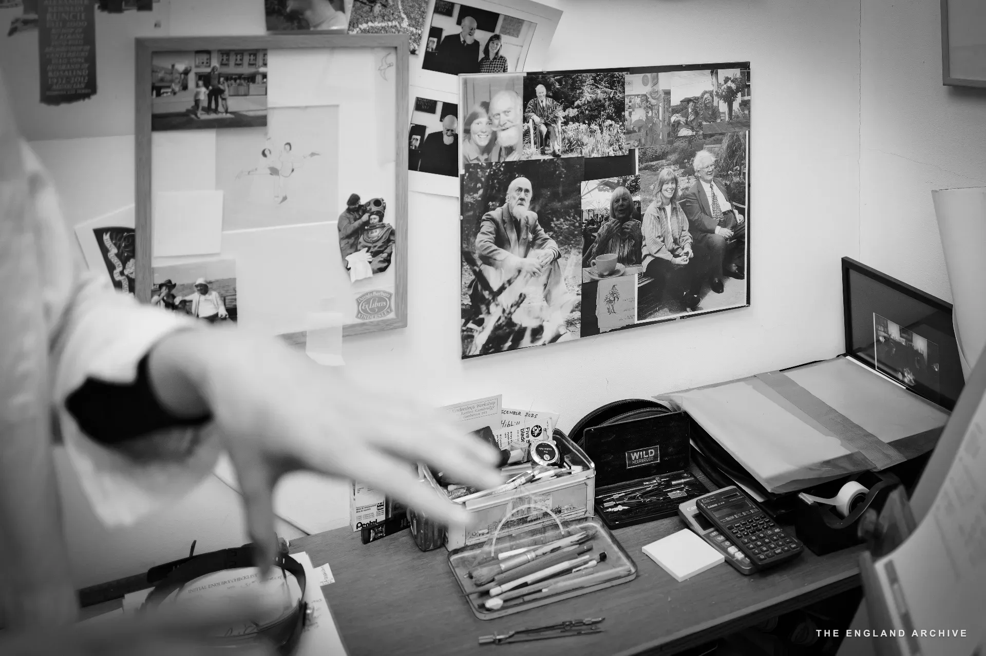 A close on the desktop in the workshop's office: a hand reaching across to a tray of pencils and pens; on the pinboard above, photographs of David Kindersley and the workshop's history; a laptop showing a video call on the right.