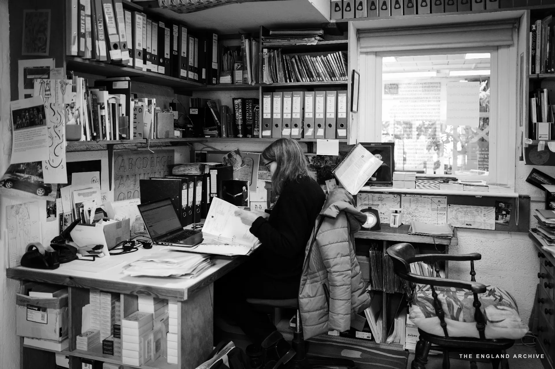 A wide view of the office: tall ranks of black-spined files filling the walls, a window letting in flat daylight, a woman at the desk under the window working with paper in her lap, a chair piled with cushions to the right.