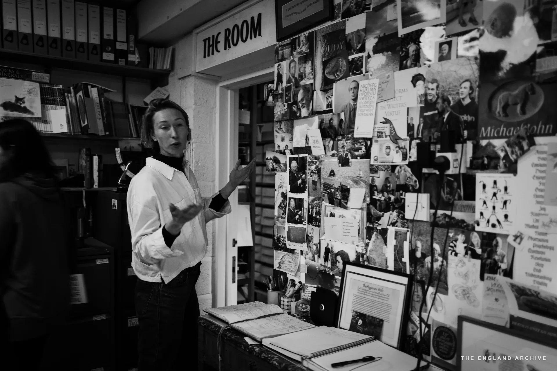 A figure in a white shirt mid-explanation gesturing at a pinboard wall covered with photographs, letters, archive material from the workshop's history. A doorway labelled 'THE ROOM' opens behind.