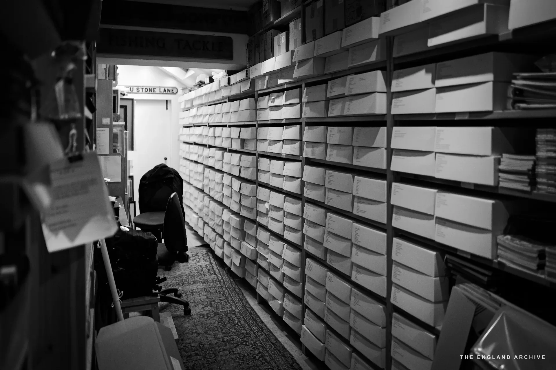 A long corridor lined floor to ceiling with white archive boxes, each labelled in hand by client name, with a person bending at the far end working on a low shelf. A high tapered light cuts down the centre.