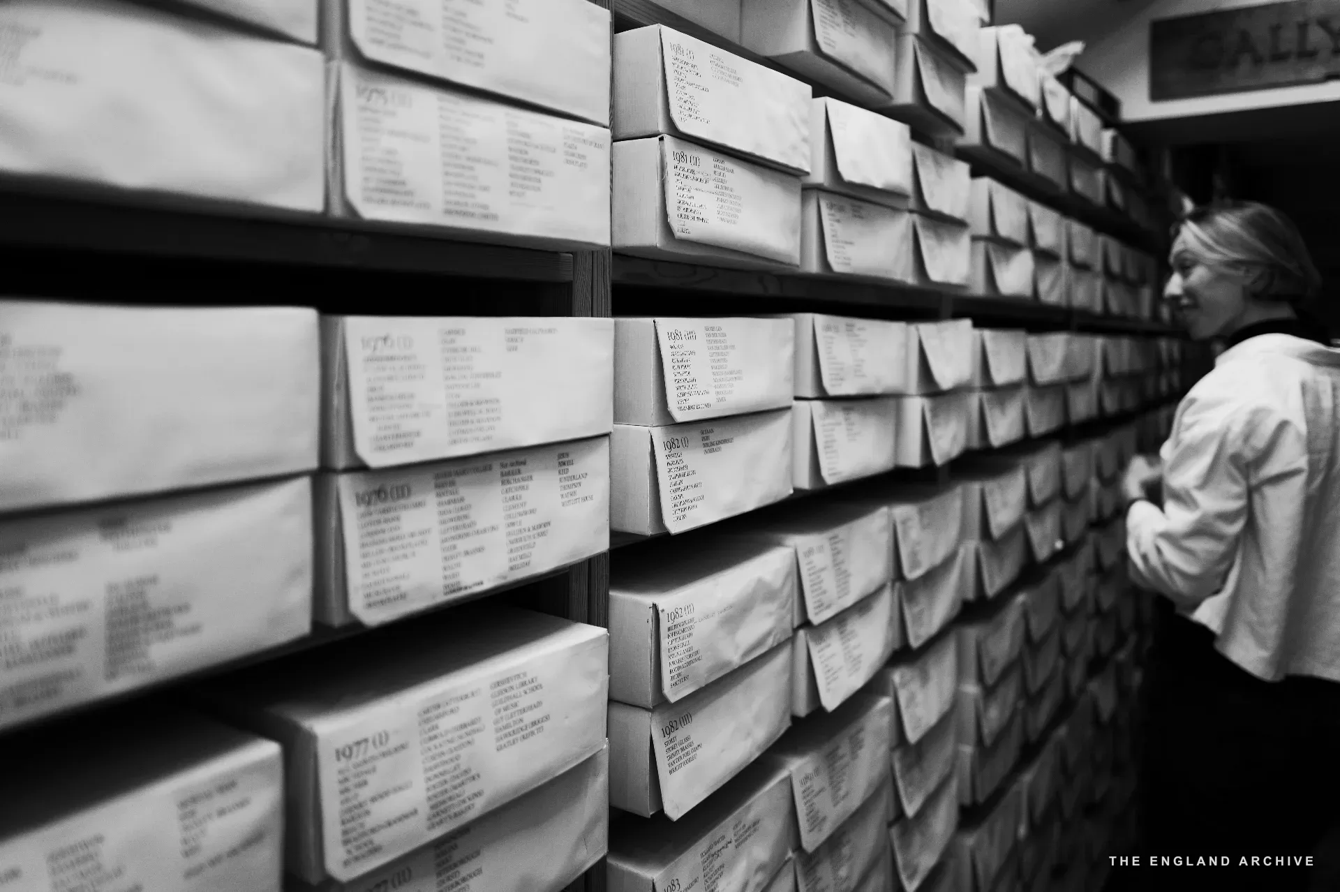 A close view of the archive box wall: dozens of white boxes with handwritten card labels visible, a worker in a white shirt to the right reaching toward one of them. Each box holds the working papers for a single commission.