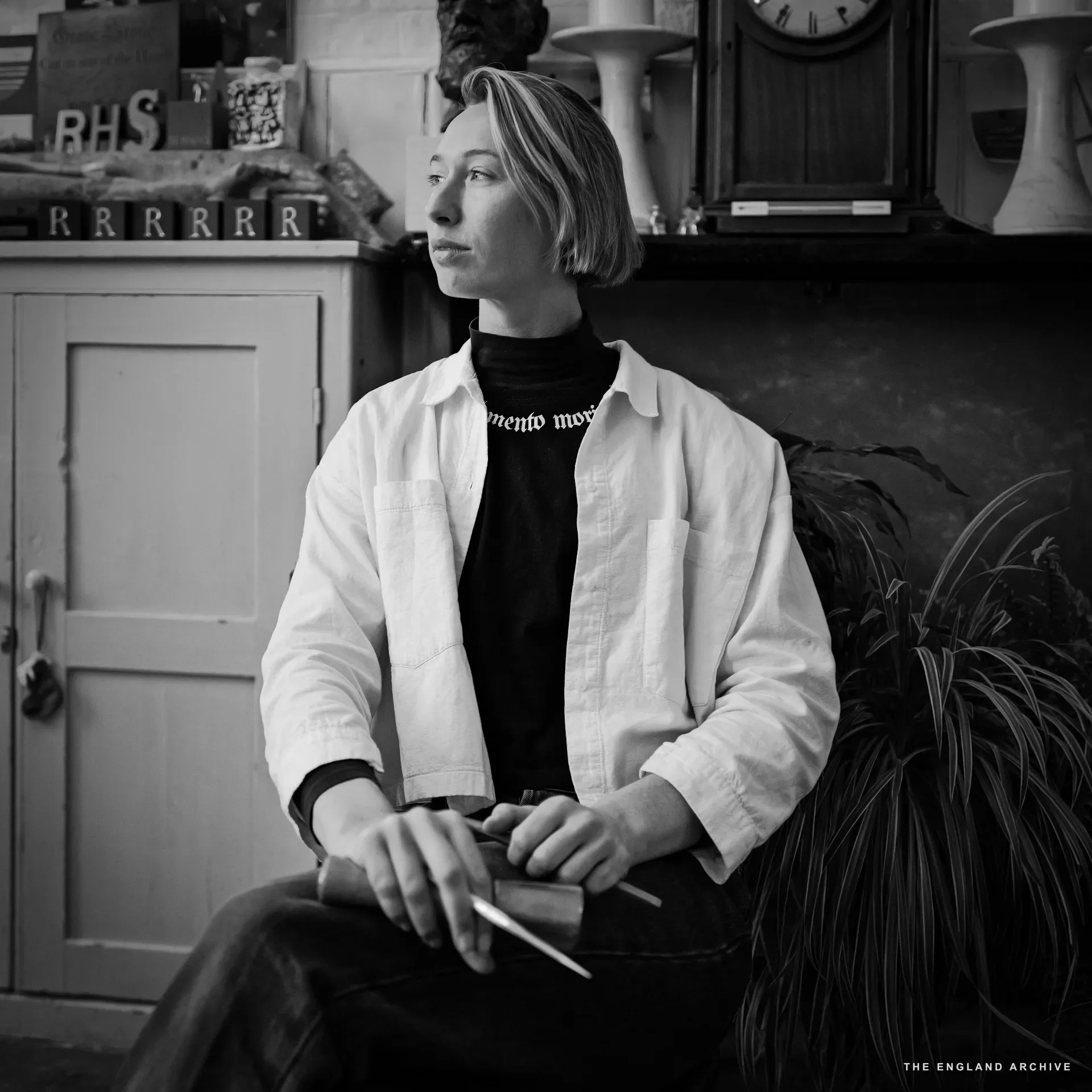 Roxanne Kindersley seated in profile for a portrait, looking off camera, in a white shirt over a 'memento mori' black T-shirt, holding a sharpened pencil and a dummy in her lap. Behind her the workshop's pigeonhole shelf, carved 'RHS' letters, the 'RRRR' alphabet row, and a longcase clock.