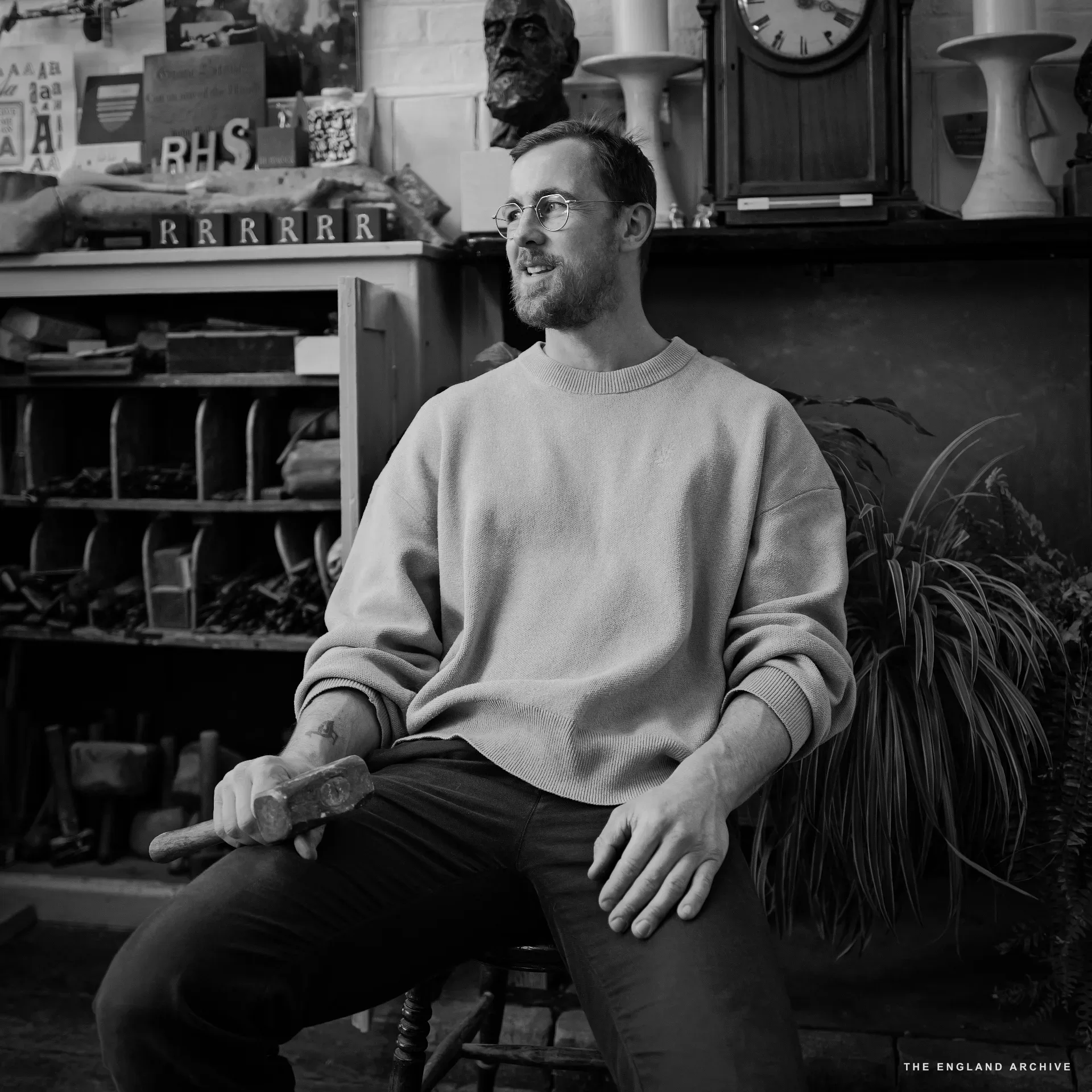 Vincent Kindersley seated for a portrait in a cream jumper, looking off camera with a faint smile, the stone-cutting hammer held loosely in his right hand. Behind him the workshop's chisel-rack pigeonholes, the 'RHS' carved letters, a bronze portrait bust, a longcase clock, and ceramic vases on a shelf.