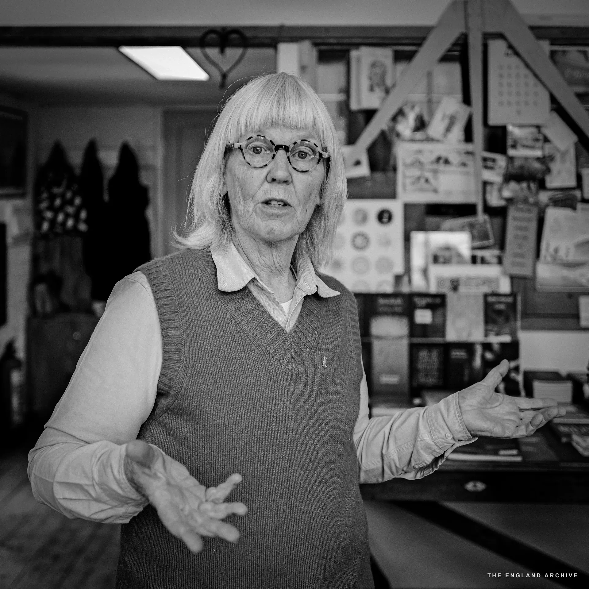Lida Kindersley standing for a portrait in her sweater vest and white shirt, both hands raised in a mid-gesture as she explains, looking past the camera. Behind her the workshop's heart-shaped iron coat-rack and the wall of pinned reference material.