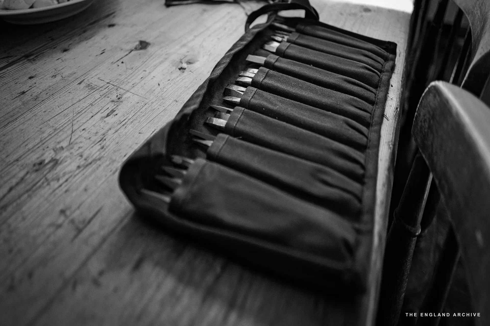 A leather chisel roll laid open on the wooden plank floor of the workshop, each pocket holding a chisel of a different size, the row of tools fading away into the depth of the frame.
