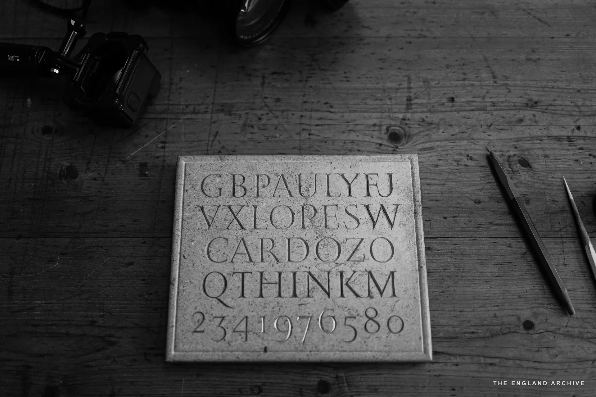 A small slate panel laid on the workshop's plank floor, cut with the workshop's lettering sample: 'GBPAULYFJ VXLOPESW CARDOZO QTHINKM 234 1976 0580'. A camera and pencils rest beside it.