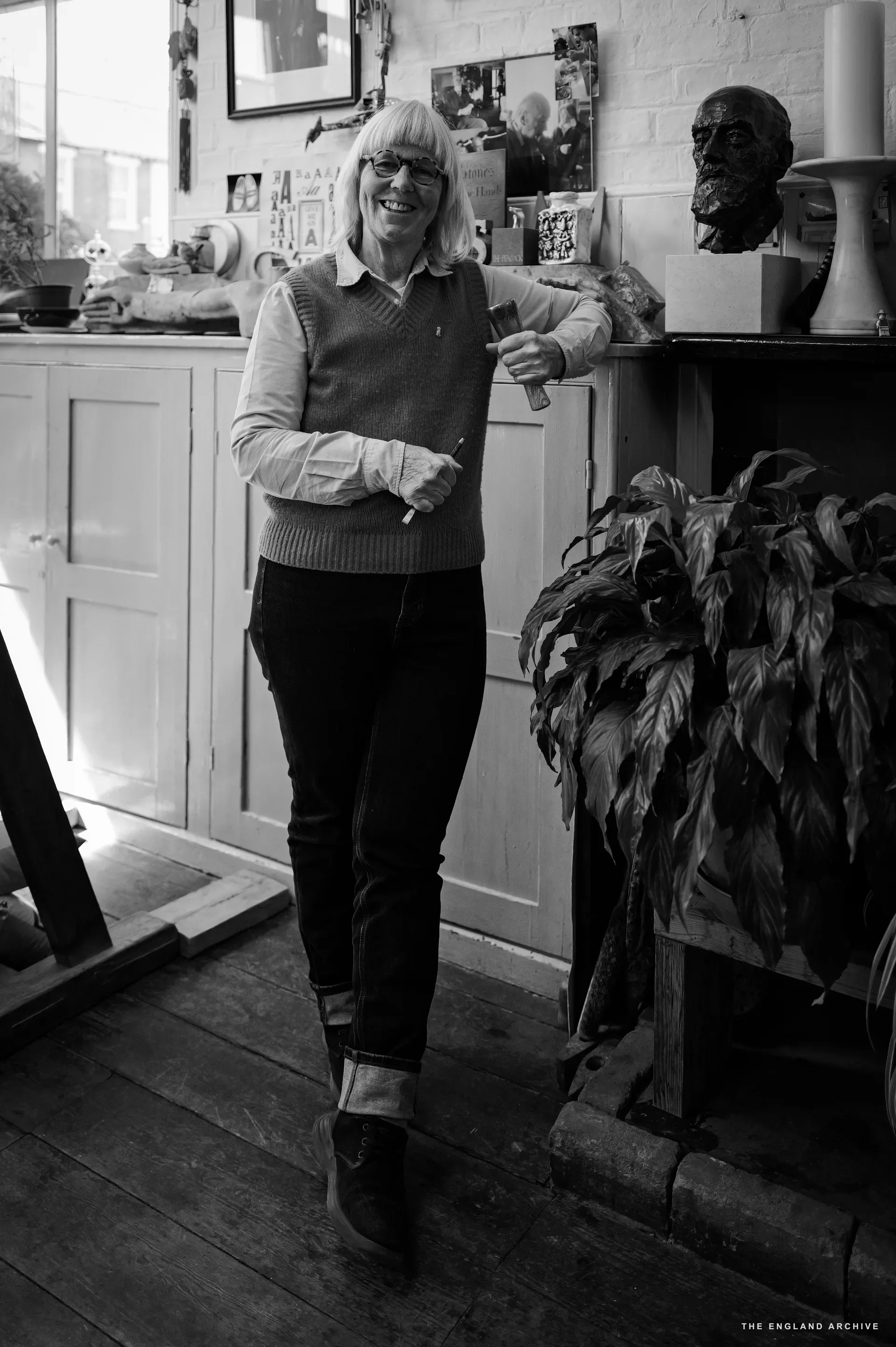 A full-length portrait of Lida Kindersley leaning casually against the kitchen counter in the workshop, smiling toward the camera, hands holding a pencil and a stone offcut. Behind her the bronze portrait bust of David Kindersley sits on a dresser; a tall houseplant fills the right side.