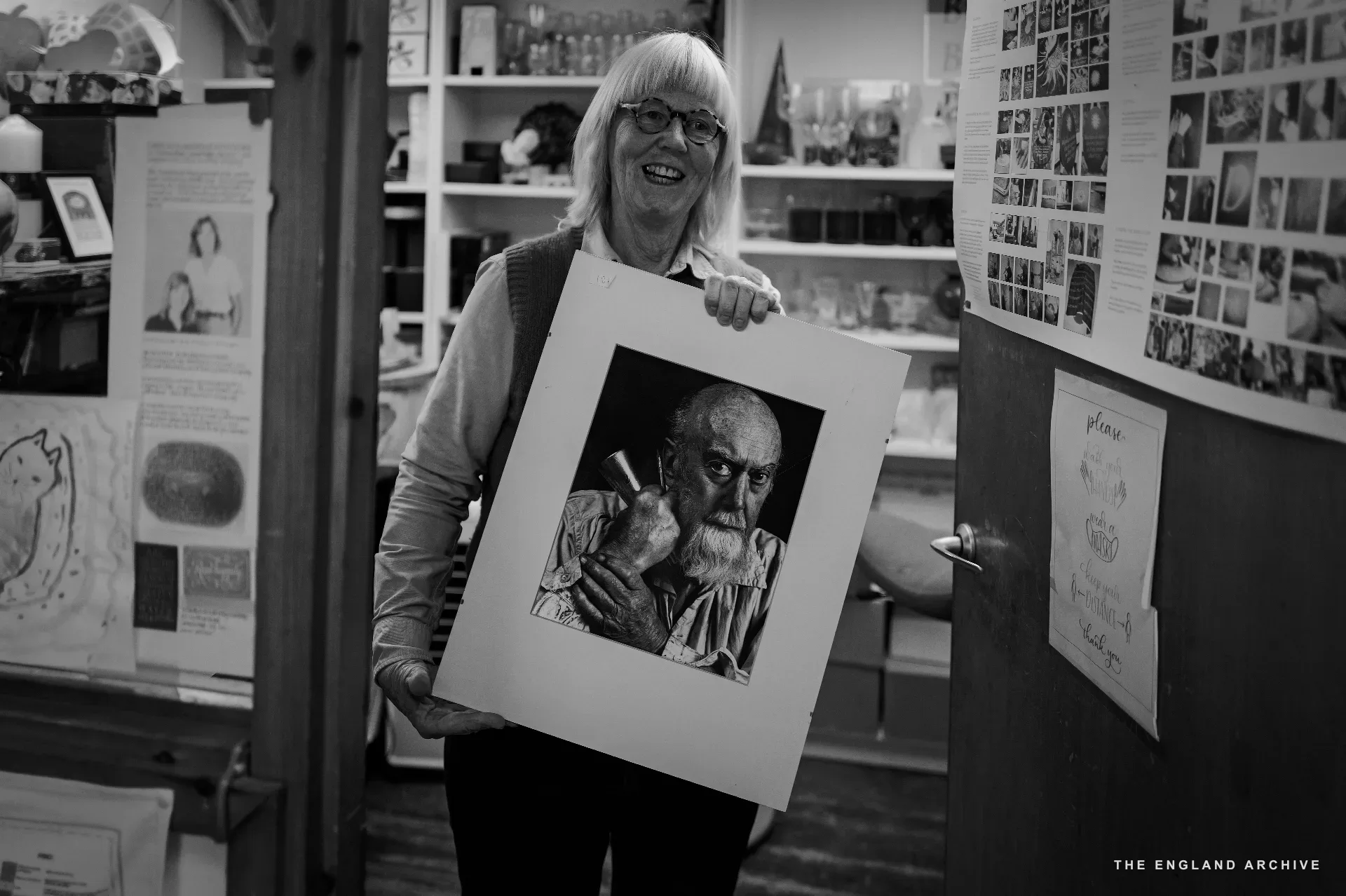 Lida Kindersley smiling, holding up a large framed black and white portrait of her late husband David Kindersley.