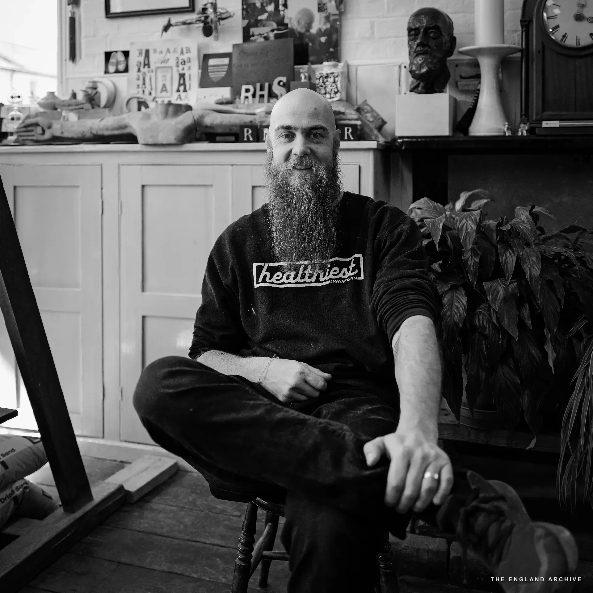 Hallam Kindersley seated for a portrait in the workshop, in a dark sweatshirt printed with the word 'healthiest', long beard, looking straight at the camera. Behind him the workshop's pigeonhole shelf, the carved 'RHS' letters, the bronze bust of his father David Kindersley, a longcase clock, and ceramic vases.