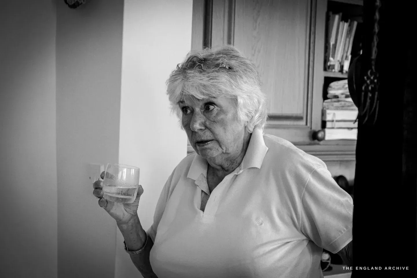 Carol standing in her kitchen mid-sentence, holding a glass of water, an oak dresser stacked with books and papers behind her.