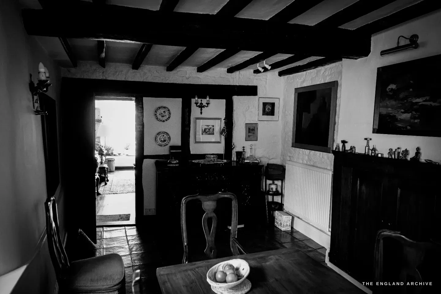 Carol's beamed dining room looking through to the sitting room beyond, dark ceiling timbers, framed seascapes and blue-and-white plates on the walls.