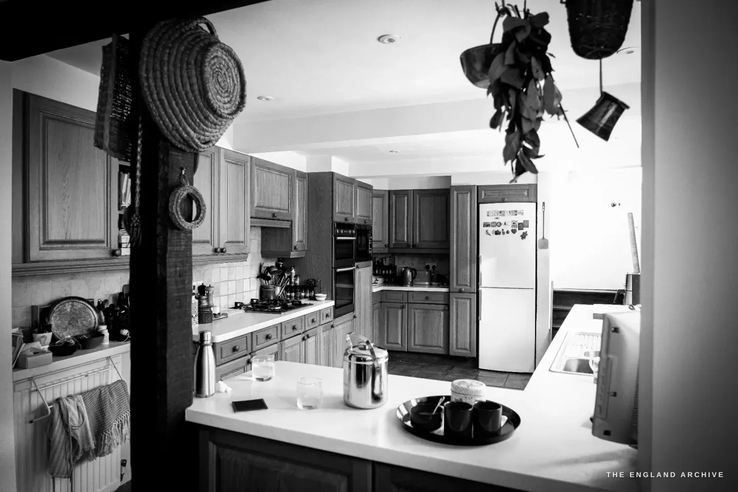 Carol's kitchen seen across the island - oak units, a tall fridge covered in magnets, baskets and dried foliage hung from a post.