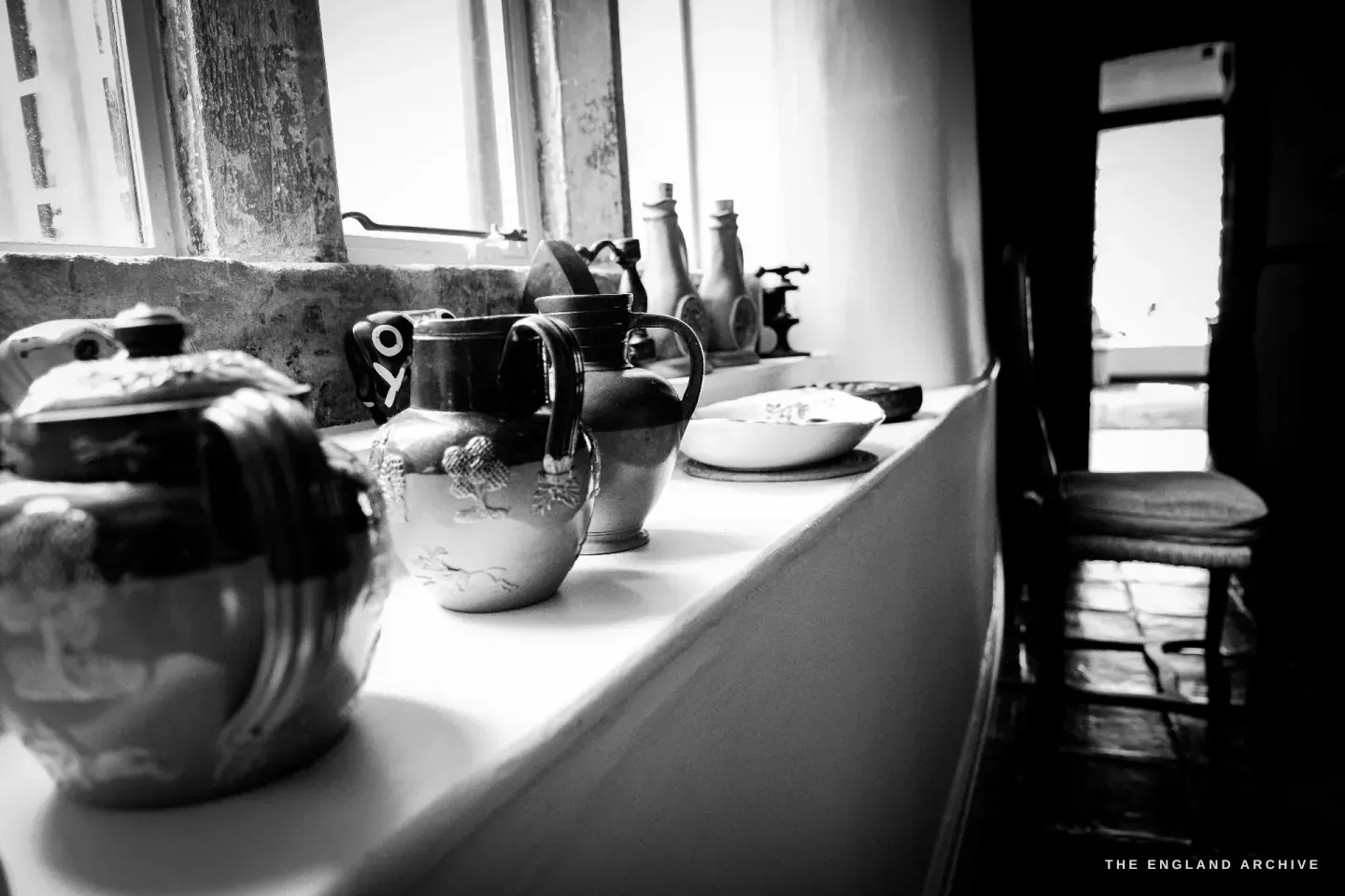 A windowsill in Carol's house lined with salt-glaze stoneware jugs, a lidded pot and pale bottles, backlit by an old painted window.