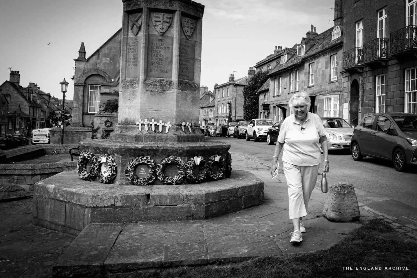 Carol walking the pavement past the war memorial and its poppy wreaths, the High Street stretching away behind.