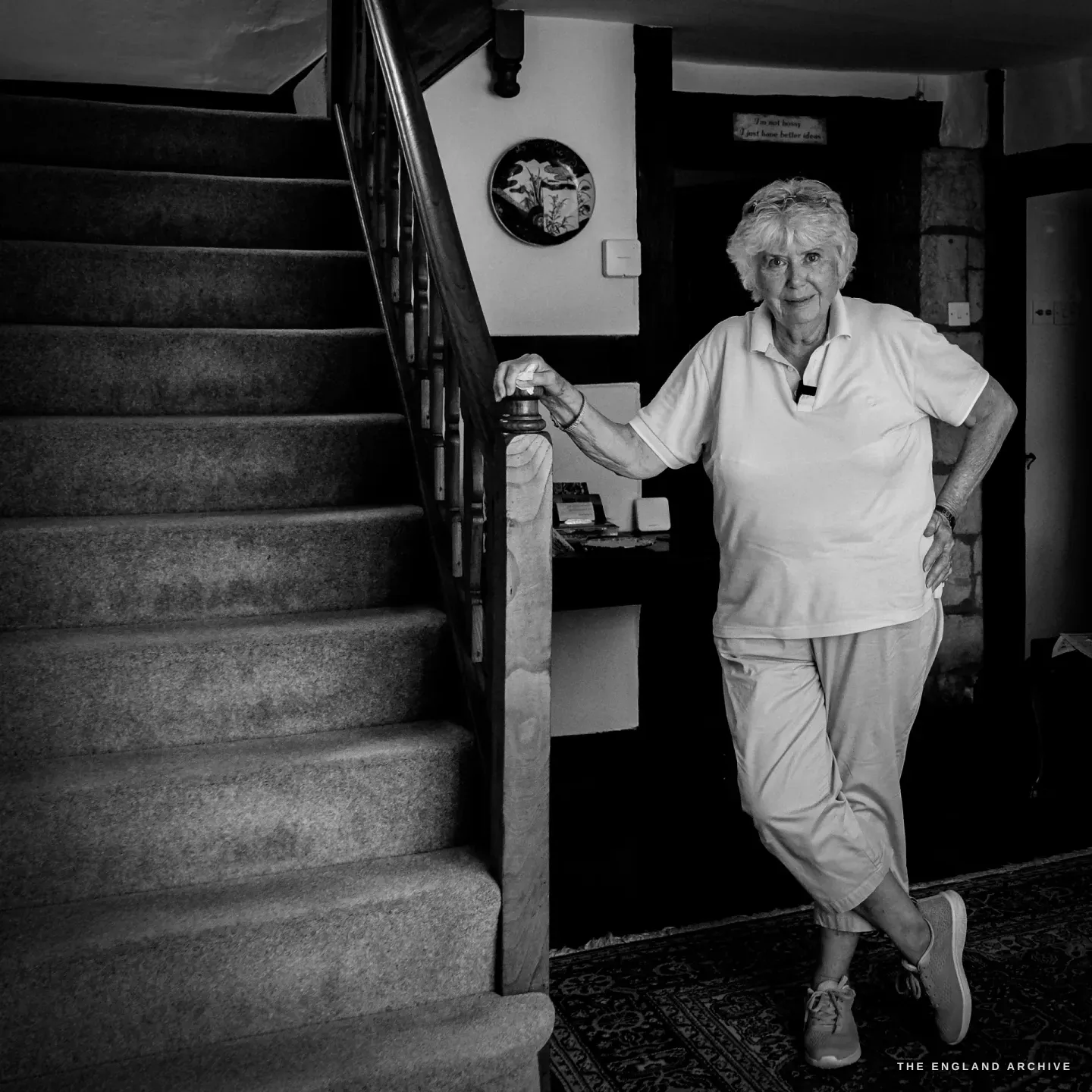 Carol at the foot of her carpeted staircase, a hand on the turned newel post, beneath a small hand-lettered house motto.