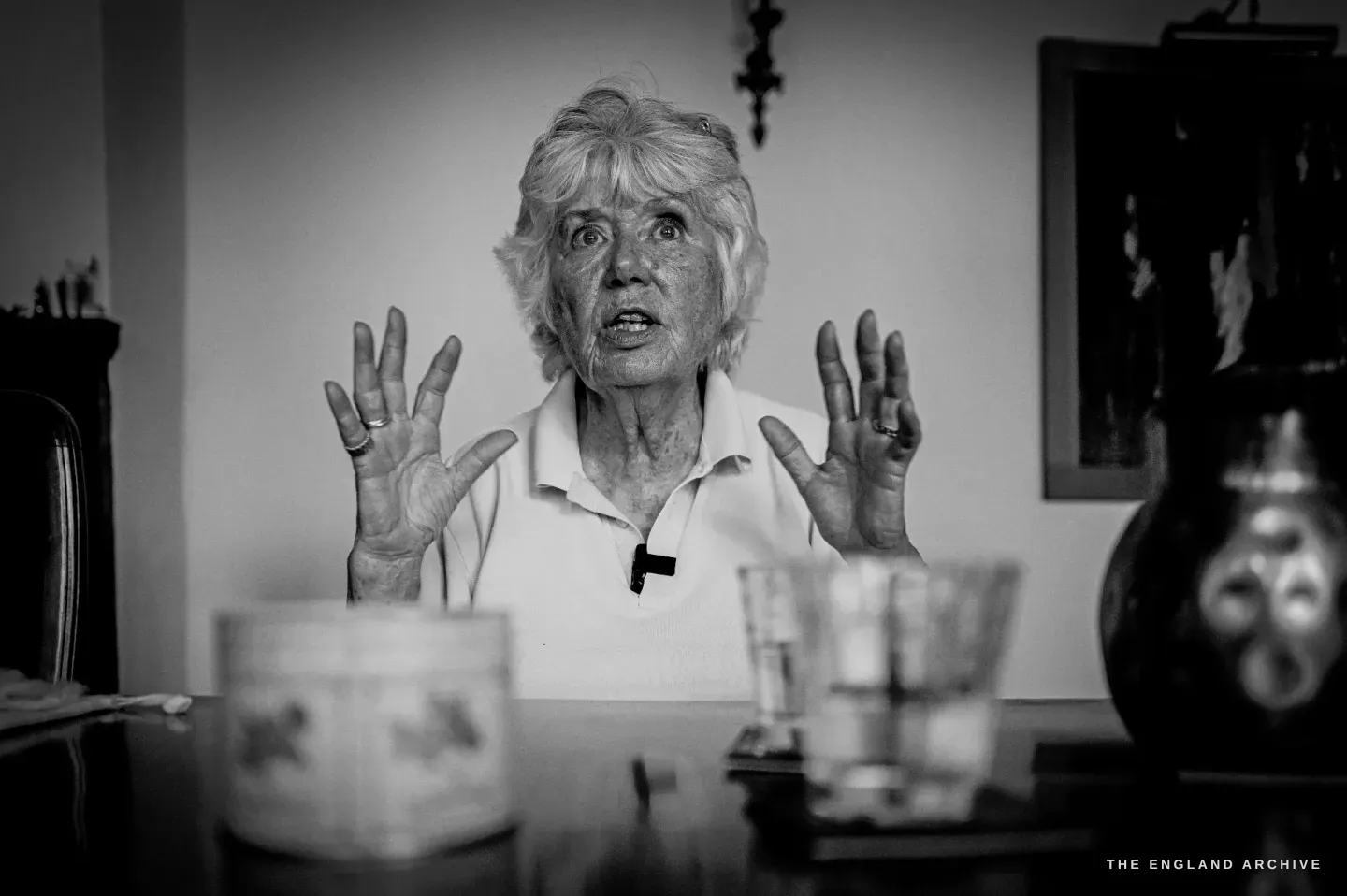 Carol at her dining table, hands raised mid-explanation, a lavalier microphone clipped to her collar and glass tumblers in the foreground.