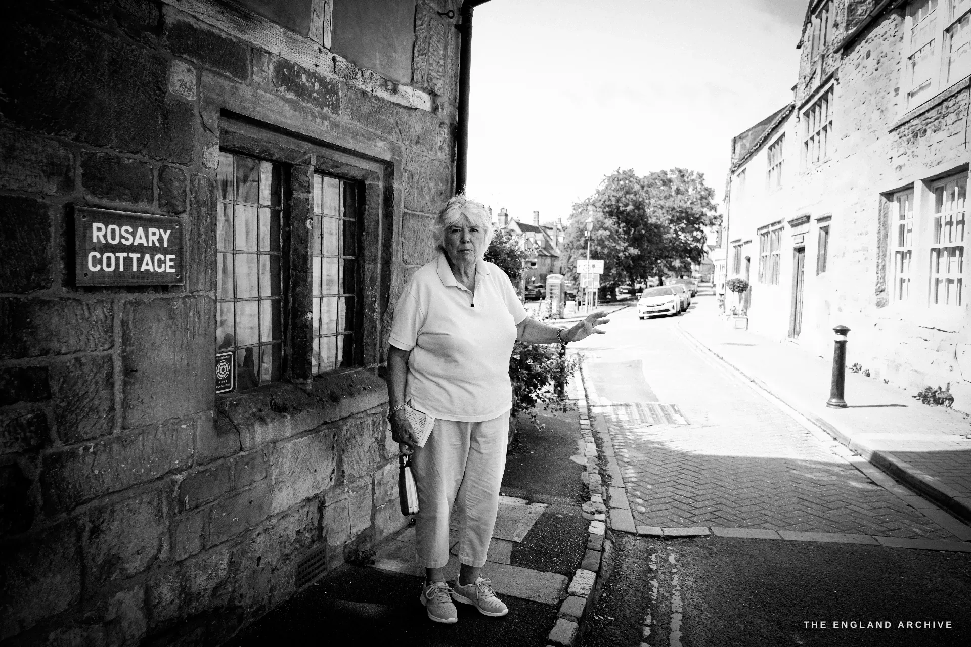 Carol stands at the corner of a stone cottage on the High Street, gesturing down the street; a red telephone box stands further along.
