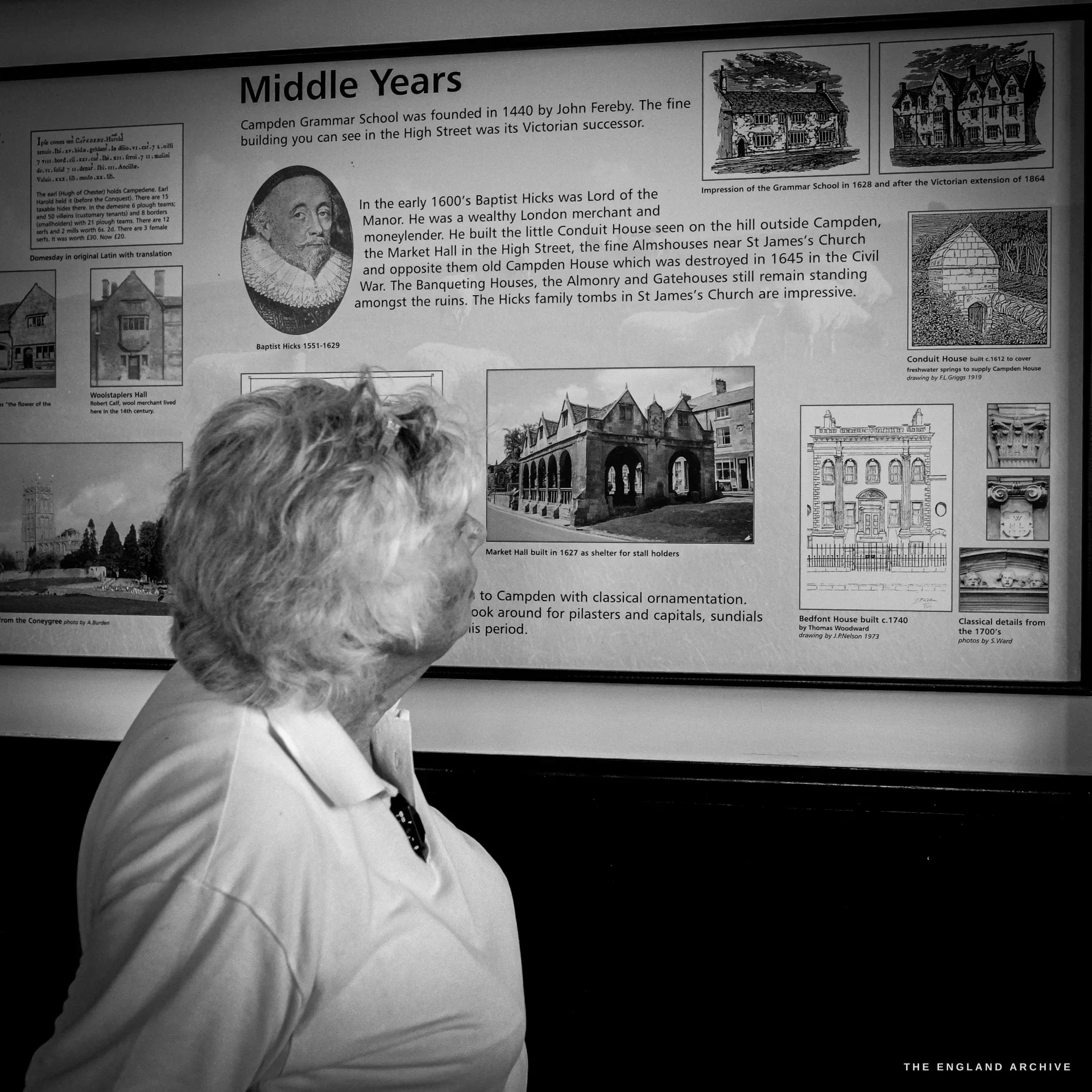 Carol stands in profile reading a framed history board titled 'Middle Years', which records the Grammar School founded in 1440 and Baptist Hicks, lord of the manor, 1551 to 1629.