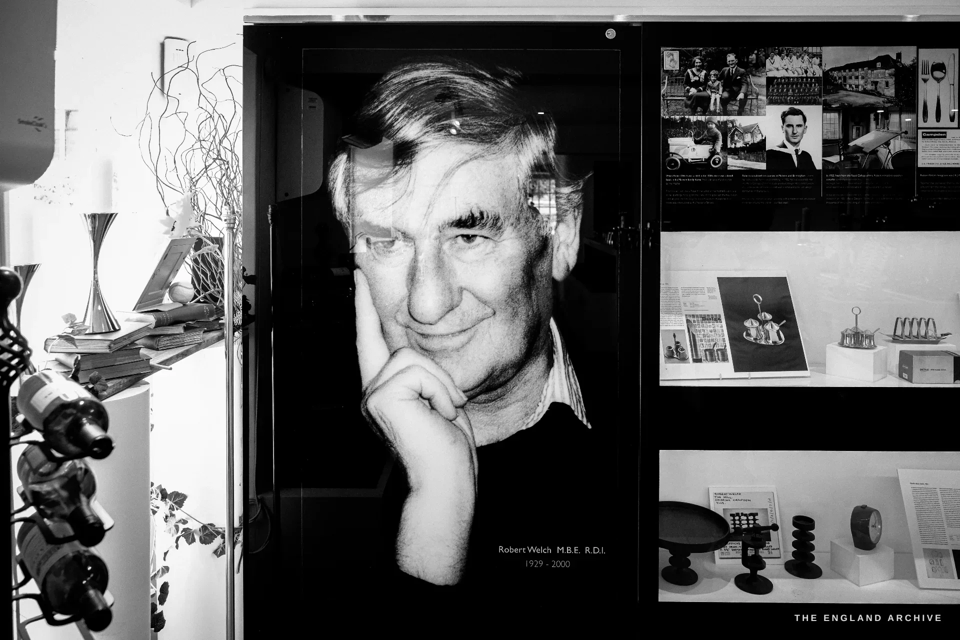 A museum display, a large backlit portrait photograph of the designer Robert Welch, hand to his chin, beside shelves of his silverware, labelled 'Robert Welch M.B.E. R.D.I. 1929-2000'.