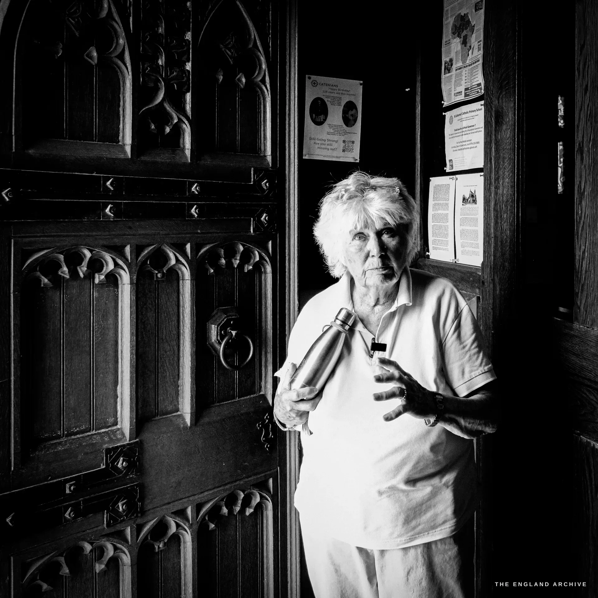 Carol stands in the dark doorway of St Catharine's beside a heavy carved Gothic oak door, holding her metal flask, a parish noticeboard behind her, looking at the camera.