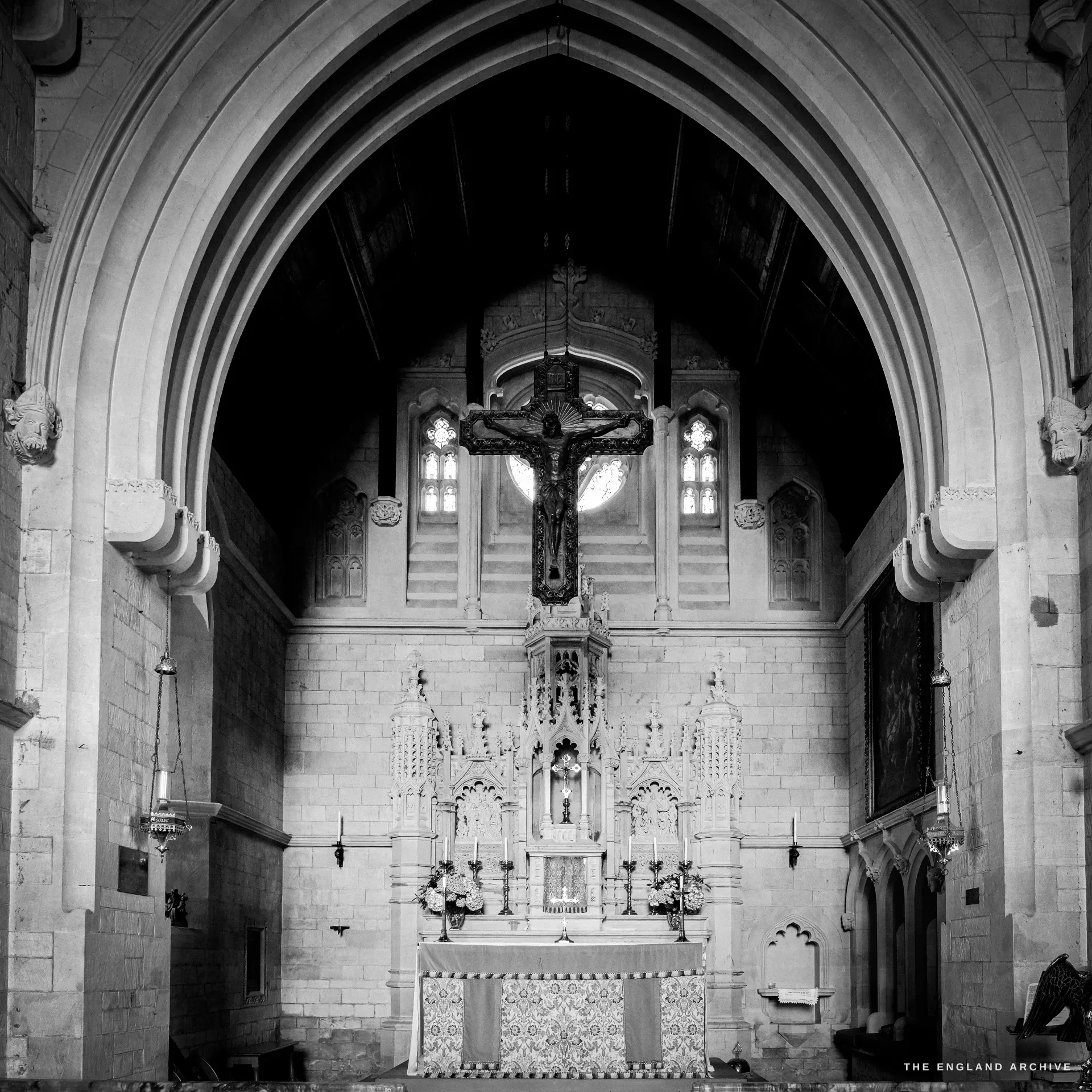 The chancel of St Catharine's Catholic Church, an ornate carved stone reredos with a crucifix suspended above it, hanging sanctuary lamps under a tall pointed chancel arch.