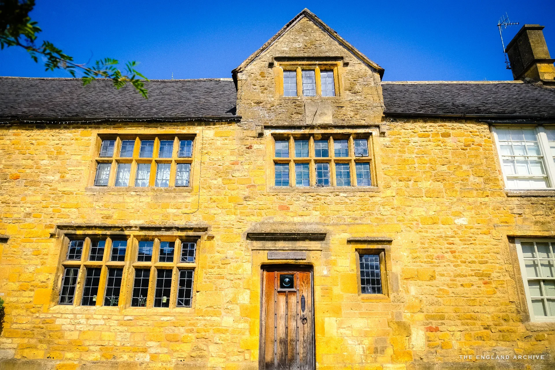 A honey-coloured Cotswold stone house front on the High Street, with mullioned and transomed windows either side of a central oak plank door.