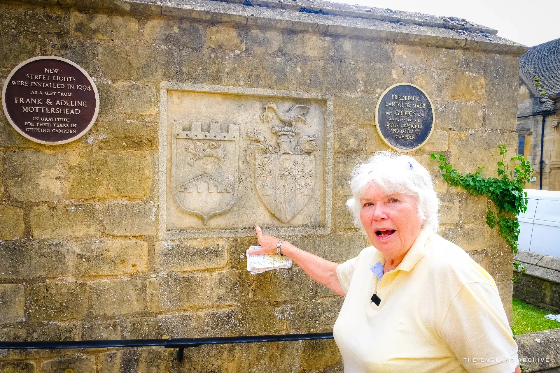 Carol points at two carved heraldic shields set into a stone wall, with two brown commemorative roundels beside them.