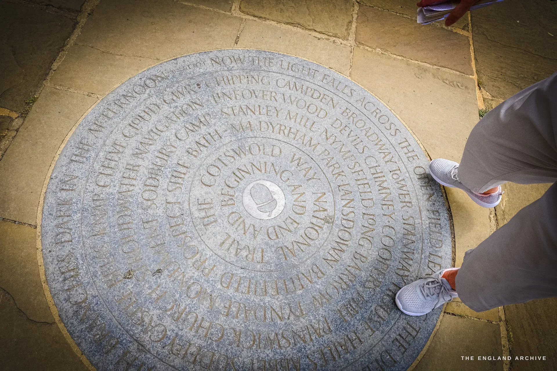 A circular carved stone pavement medallion marking the Cotswold Way, its spiralling inscription naming the stages from Chipping Campden to Bath, a pair of feet beside it.