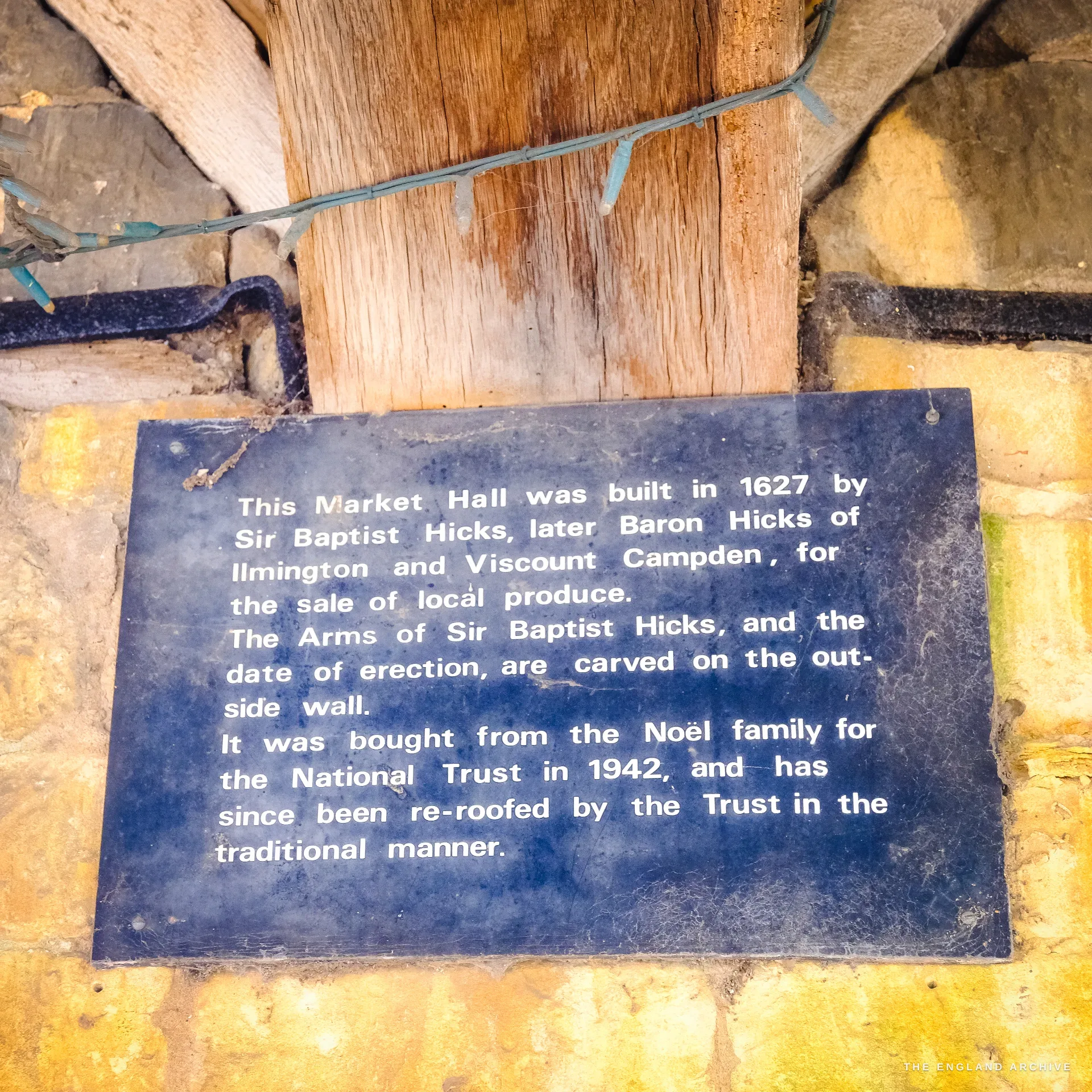 A dark cast information plaque on a timber post of the Market Hall, recording that Sir Baptist Hicks built it in 1627 and that the National Trust bought it in 1942.