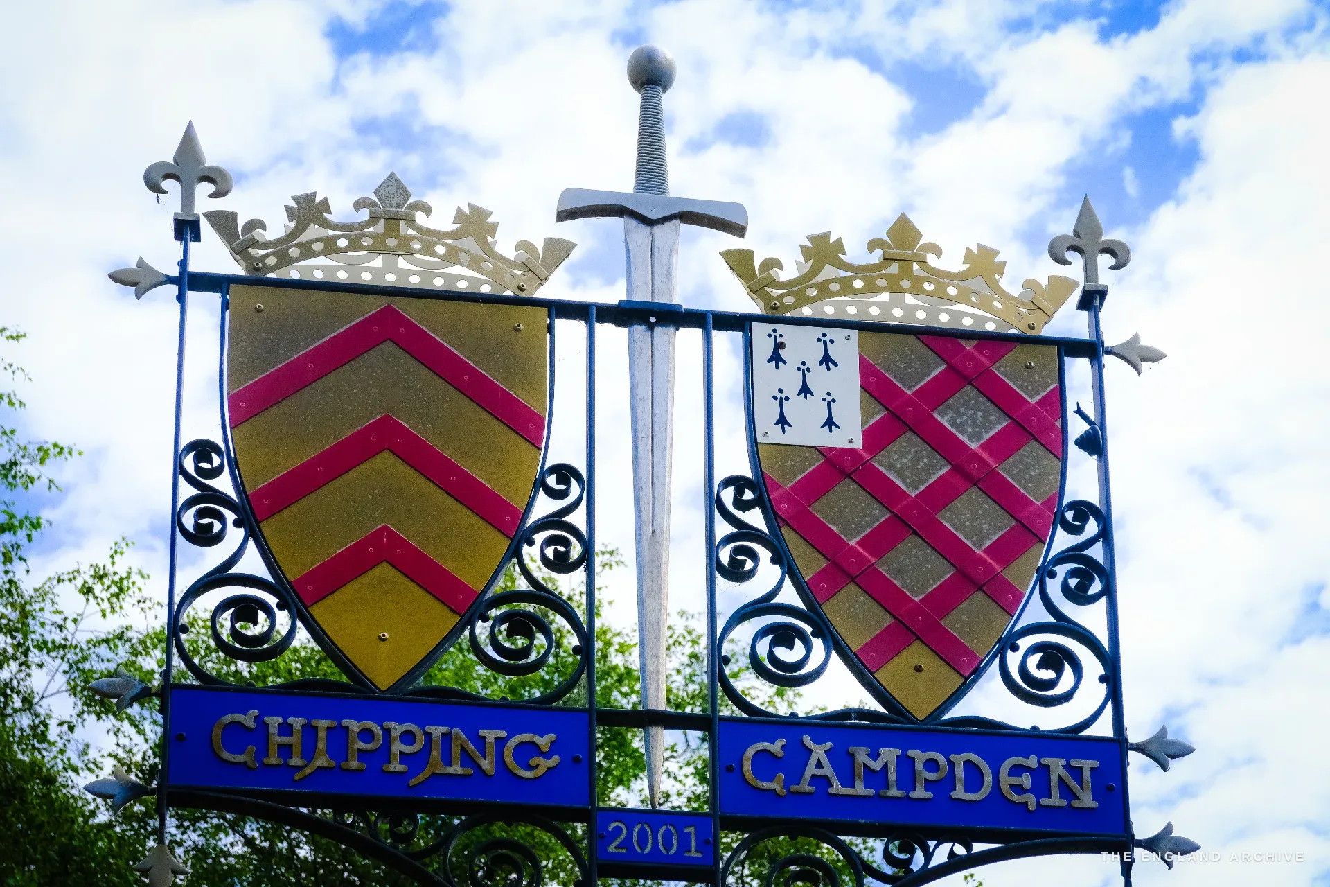 Close on the ornamental village sign - two crowned heraldic shields flanking a sword, lettered 'Chipping Campden' and dated 2001.